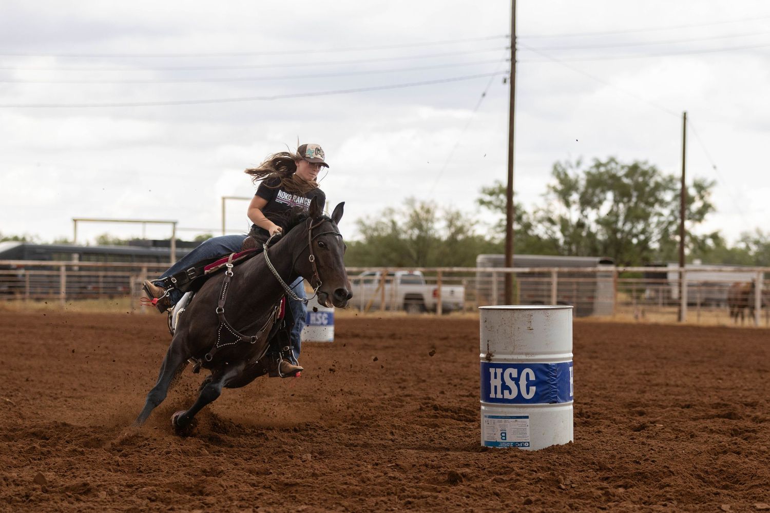 A cowgirl rides a black horse around an HSC barrel during a competitive barrel racing event at a dirt rodeo arena.