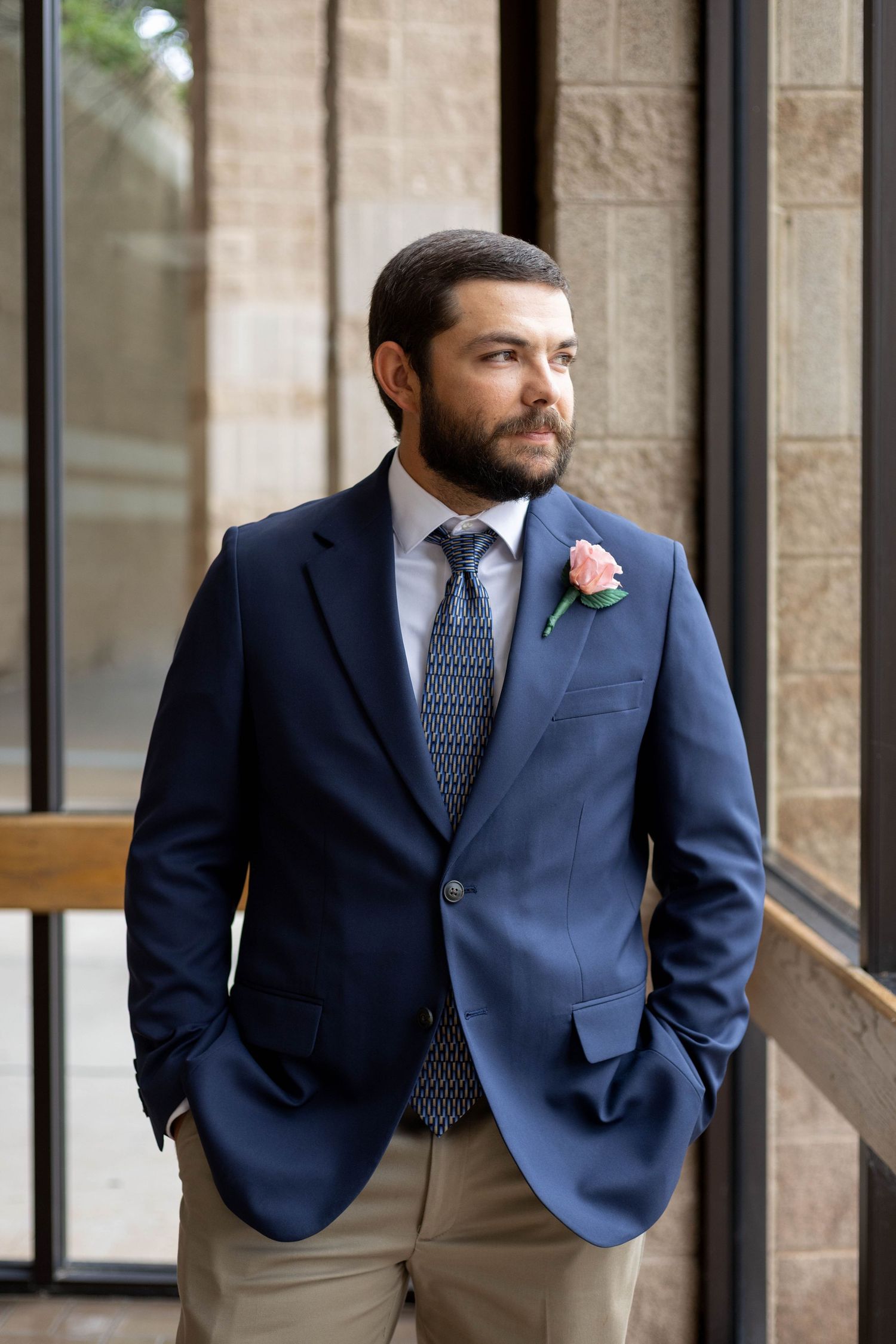 Bearded groom in navy blazer with pink boutonniere standing by stone wall.