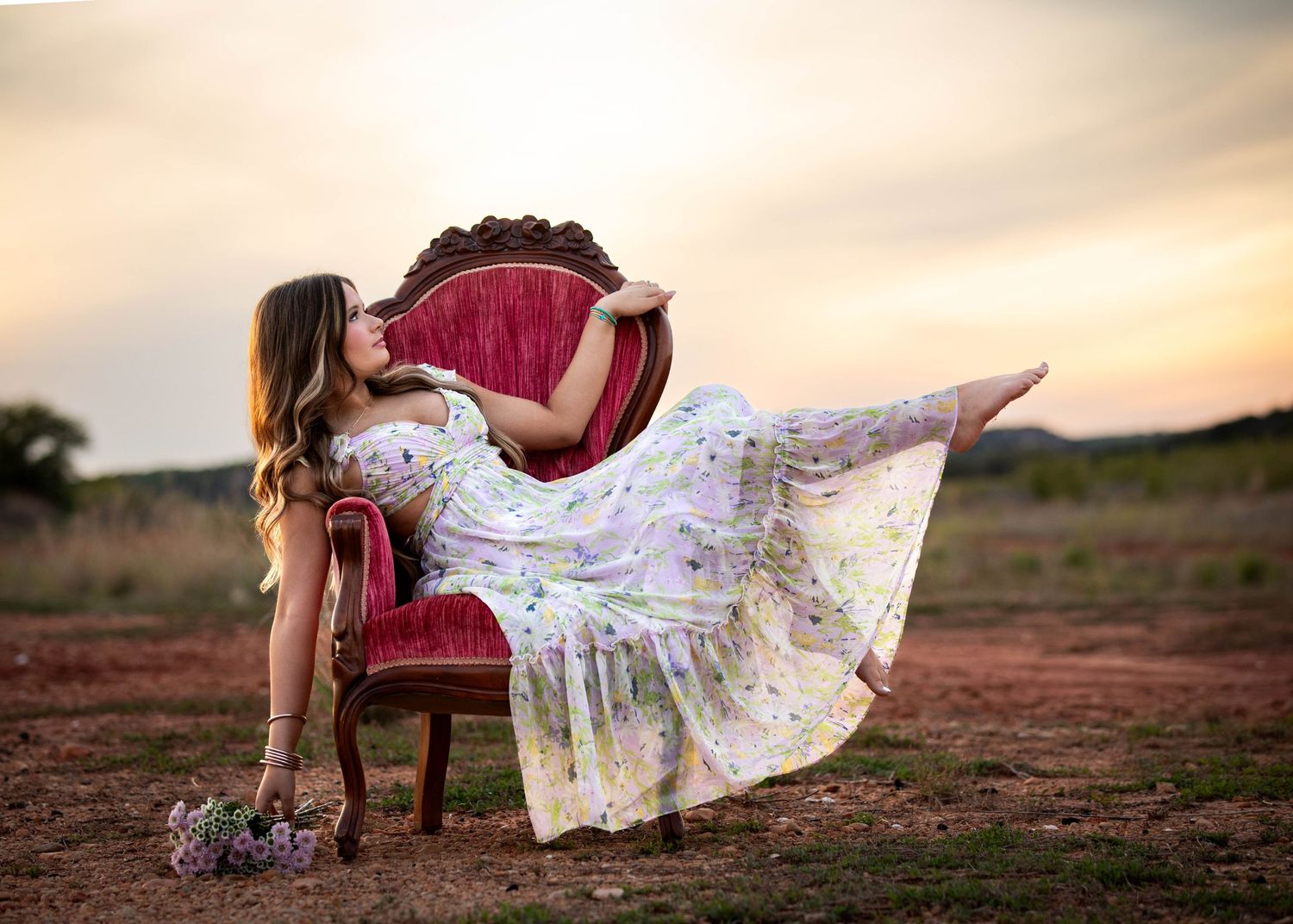 Young woman in floral dress posing dramatically in a red velvet chair in an open field at sunset.