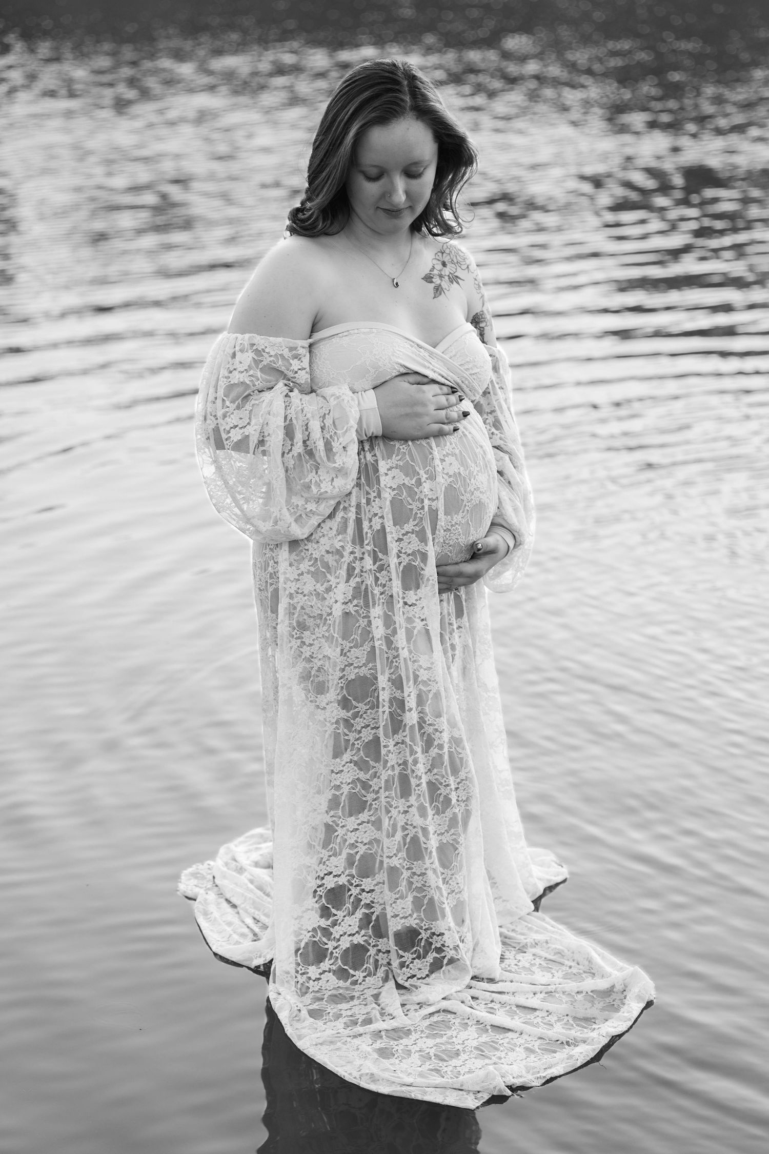 Black and white maternity photo of pregnant woman in lace gown standing in water, cradling her baby bump.