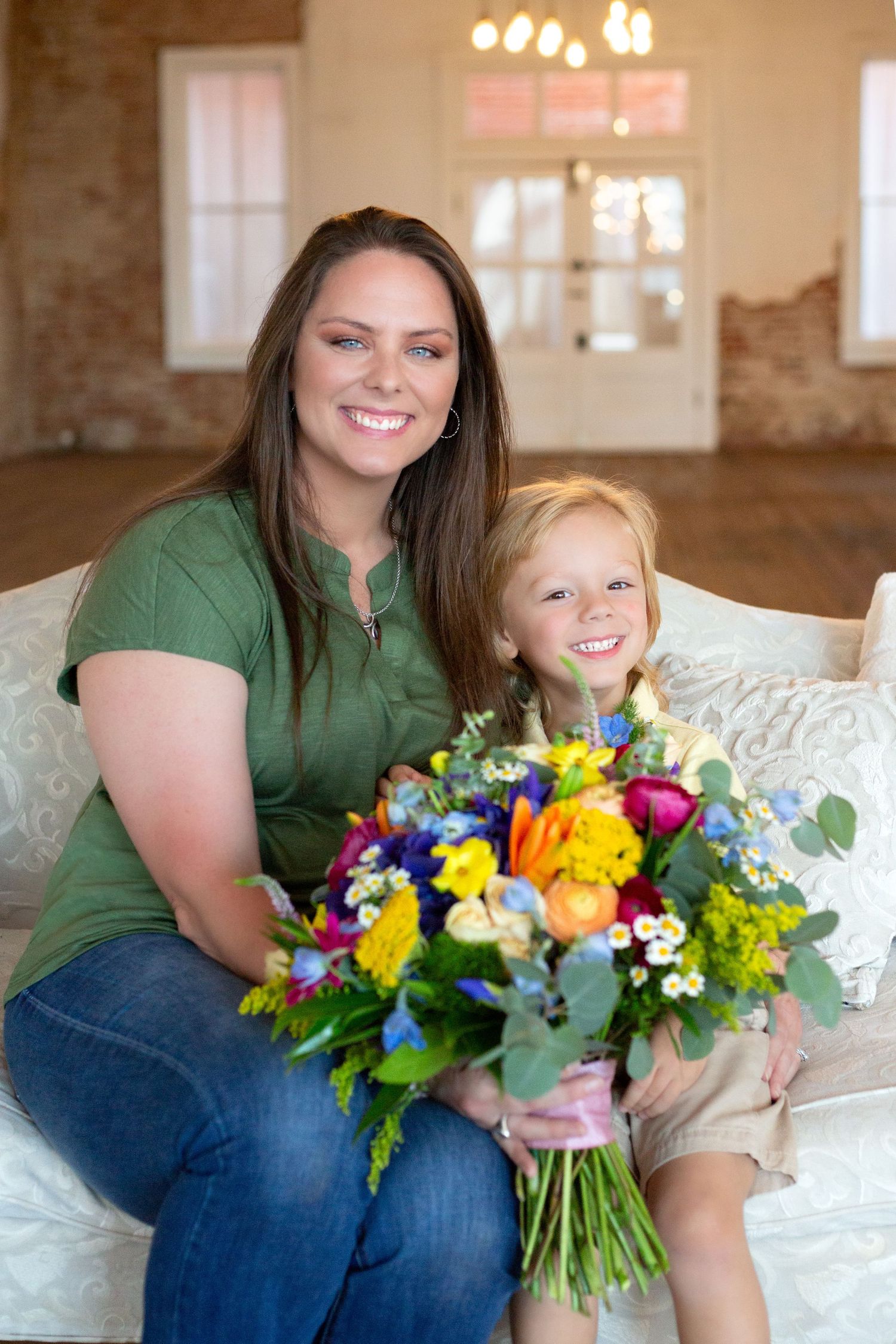 Smiling woman in green shirt sitting with young child holding a colorful flower bouquet indoors.