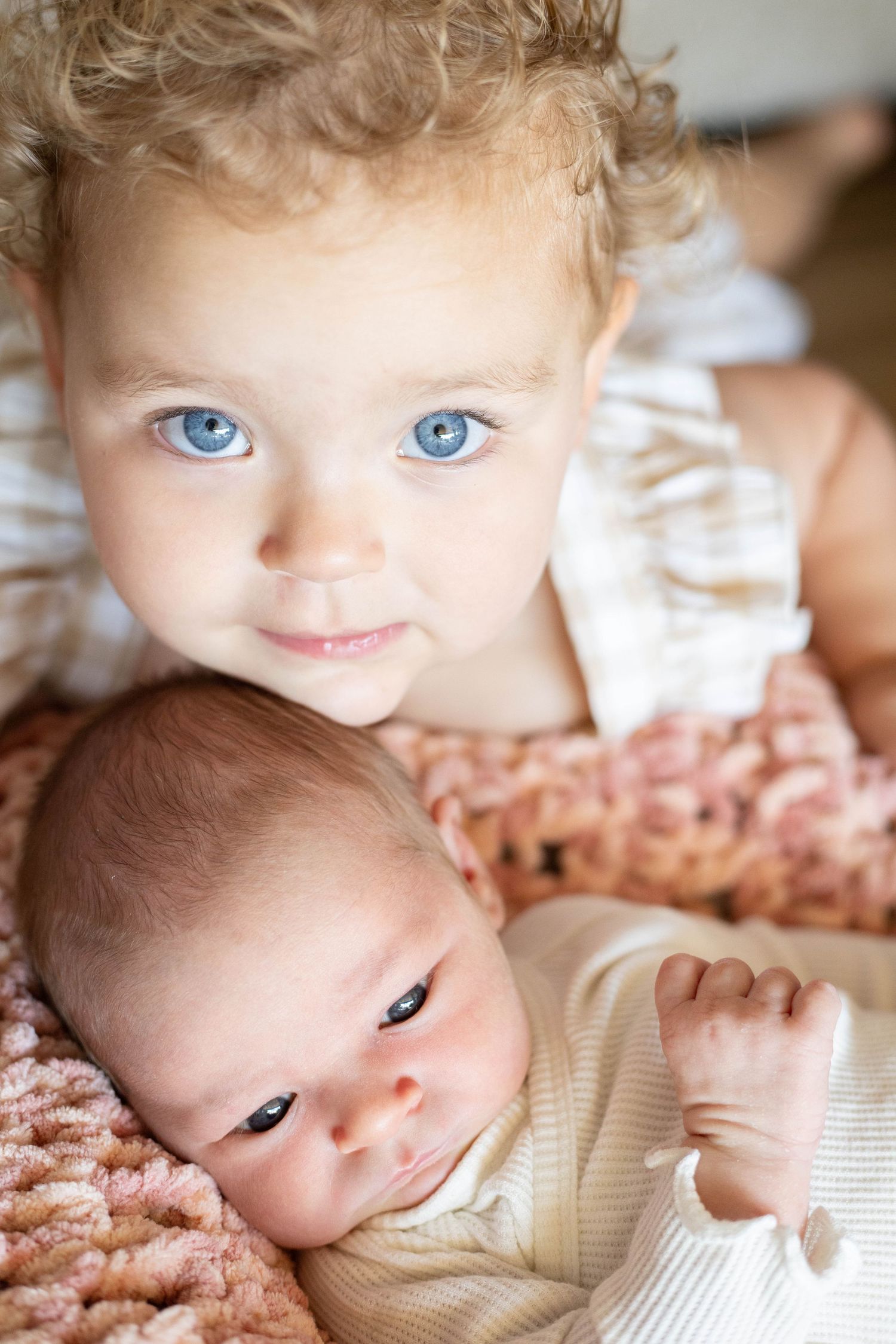 Toddler with blue eyes holding a newborn baby on a pink knit blanket.