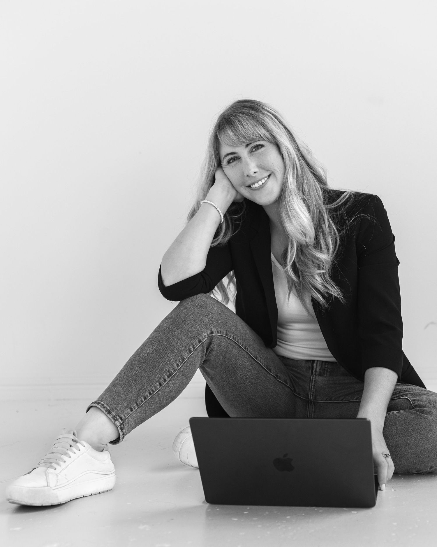 Smiling blonde woman in black blazer sitting on floor with Apple laptop in black and white photo.