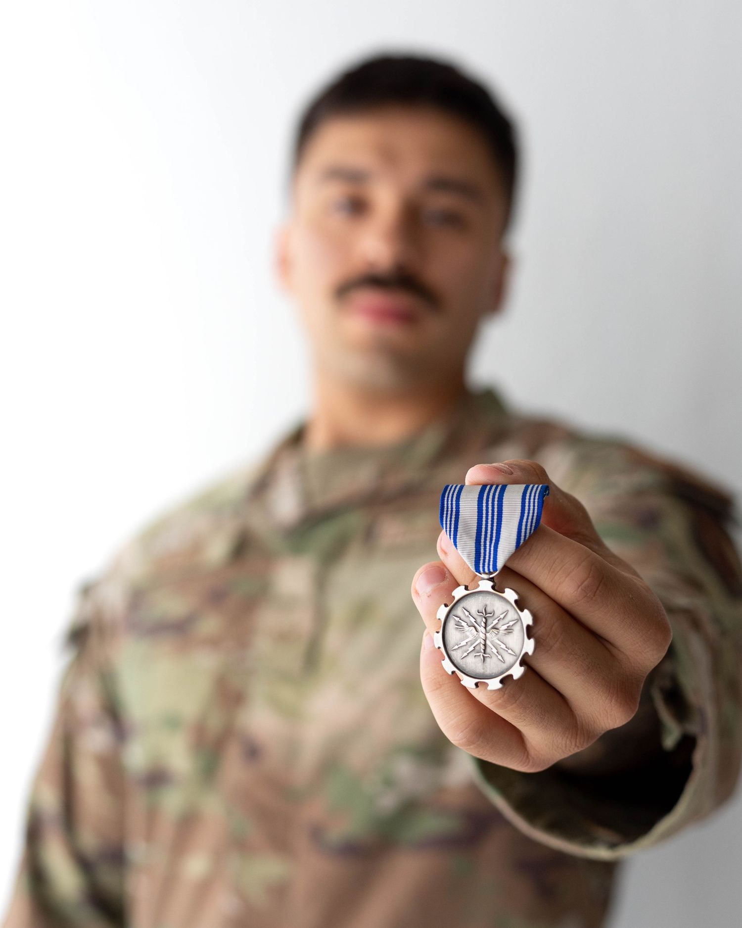 Military soldier in camouflage uniform proudly holding an achievement medal with blue ribbon toward camera.