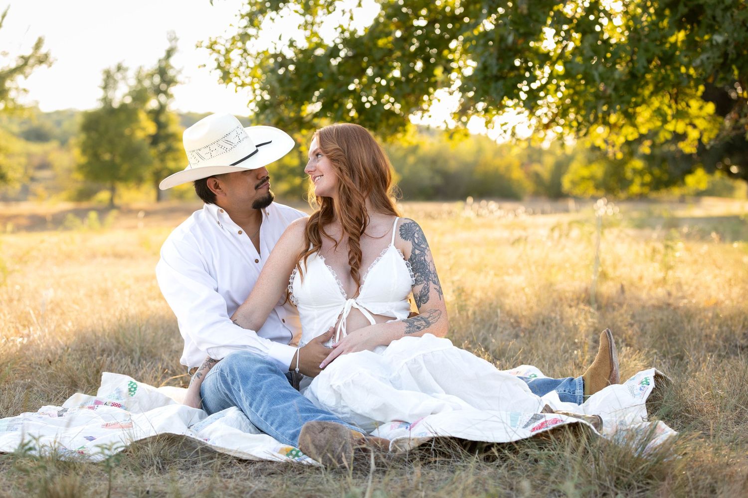 Couple sitting on a blanket in a golden field, man in cowboy hat and woman in white dress sharing a romantic moment.