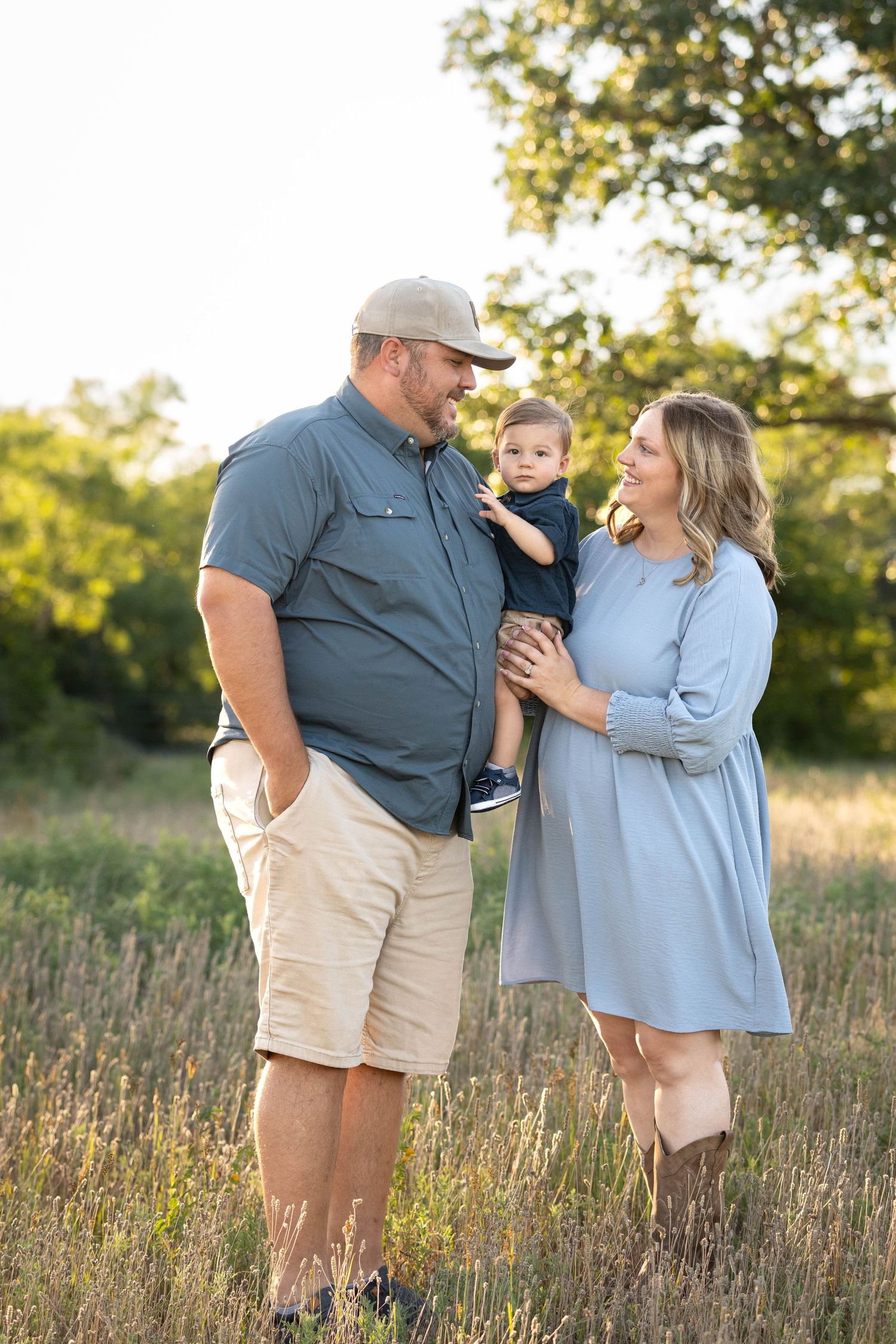 Couple smiling at each other while holding their baby boy in a golden sunlit field.