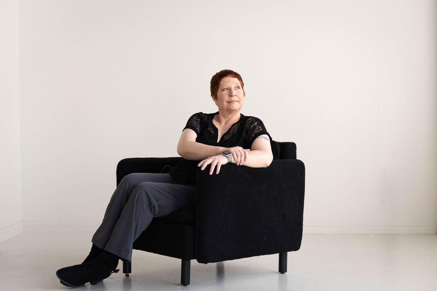Woman in black outfit sitting relaxed in a black chair against a white background.