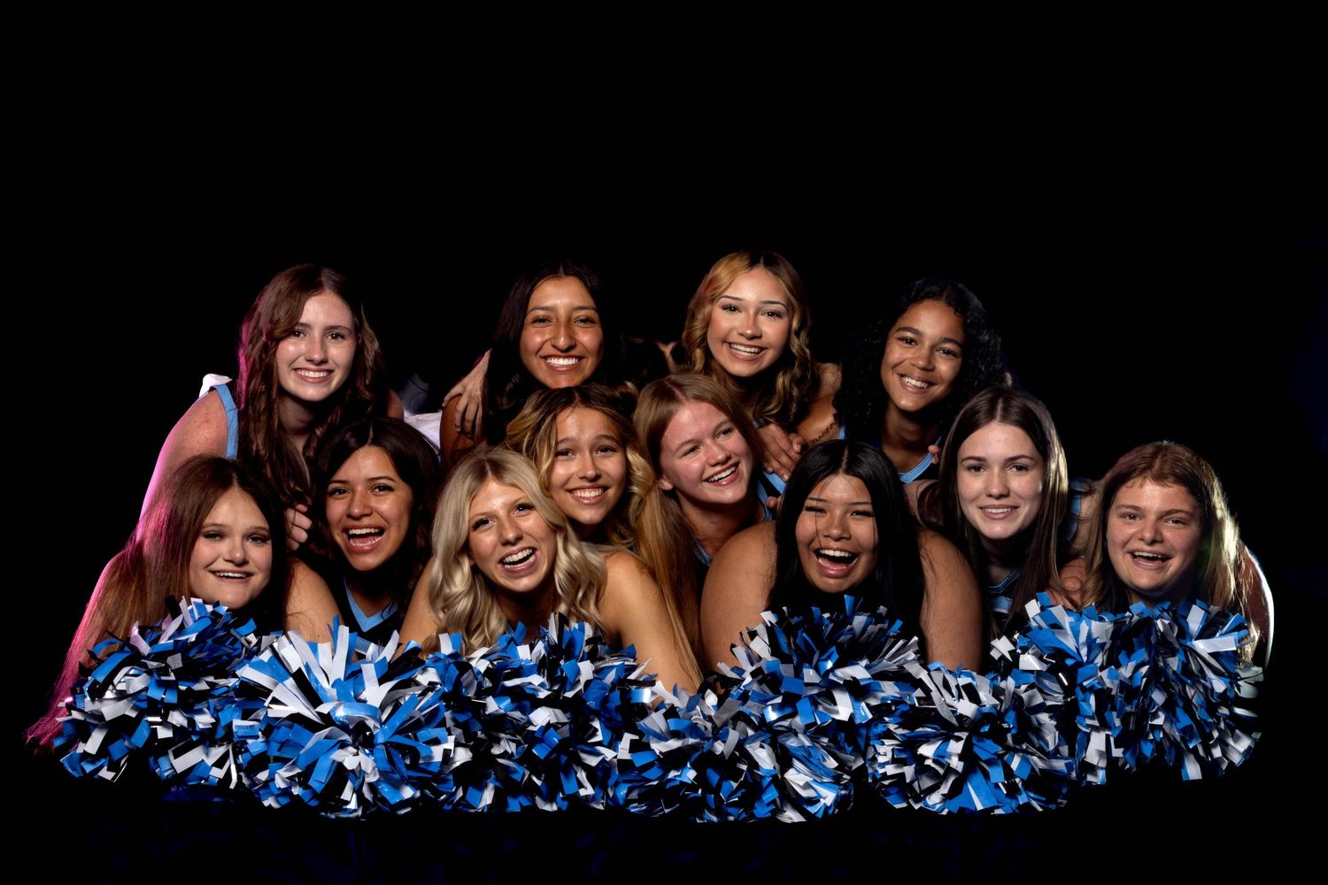 Group of cheerleaders smiling and posing with blue and silver pom-poms against a dark background.