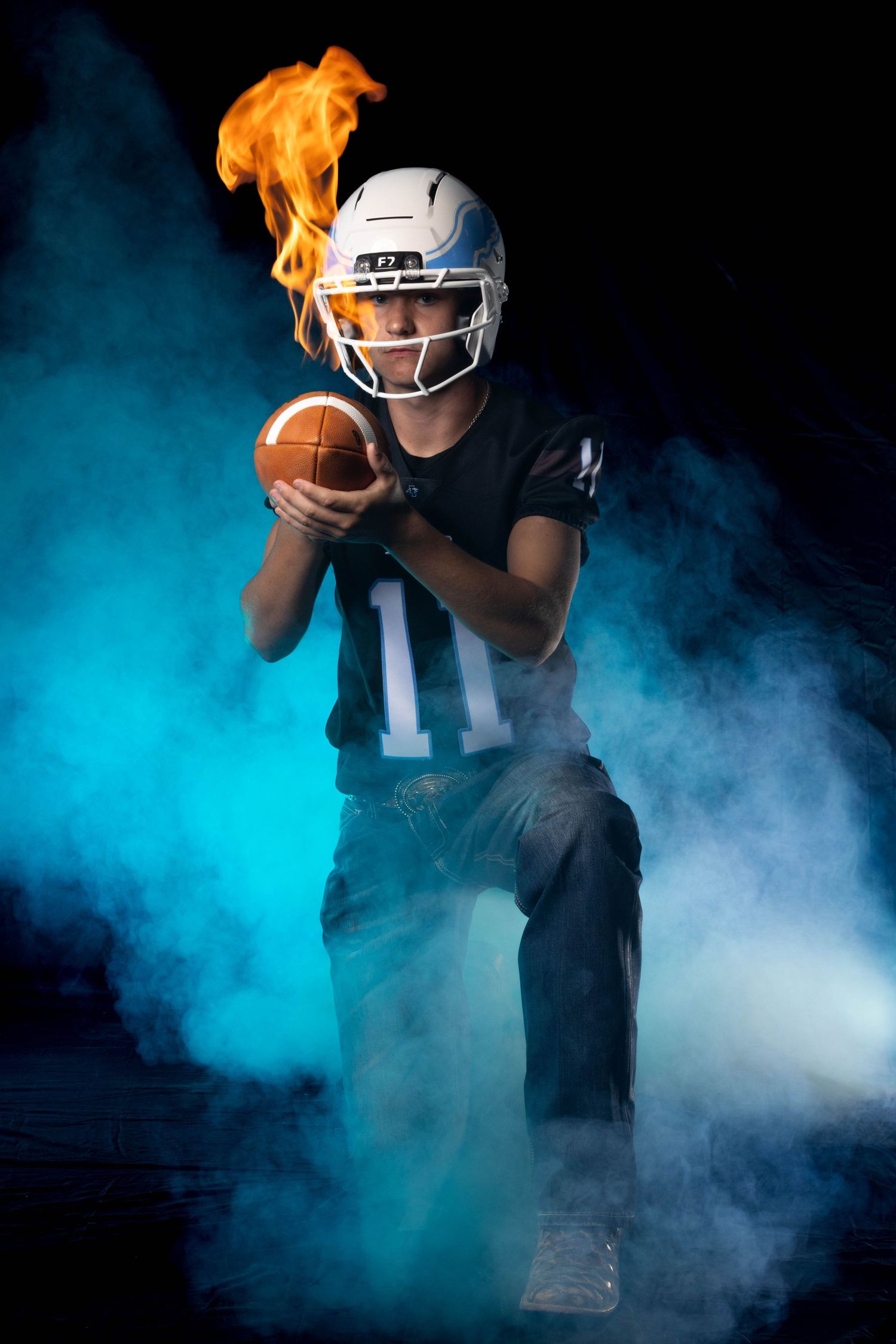 Young football player in white helmet holds a flaming ball amid blue smoke in a dramatic studio portrait.