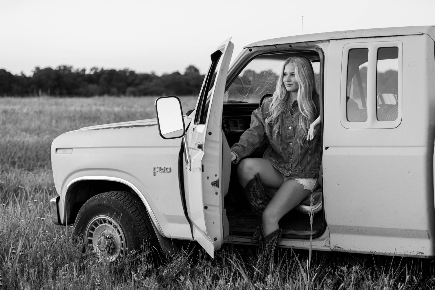 Black and white photo of a blonde woman stepping out of a vintage white truck in a grassy field.