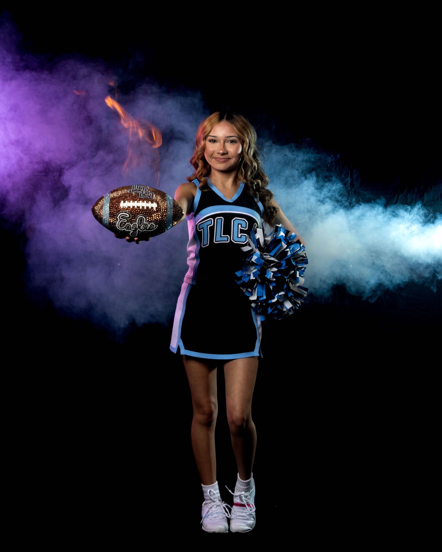 Cheerleader in TLC blue camo uniform holds football and pom-pom against dramatic purple and blue smoke background.