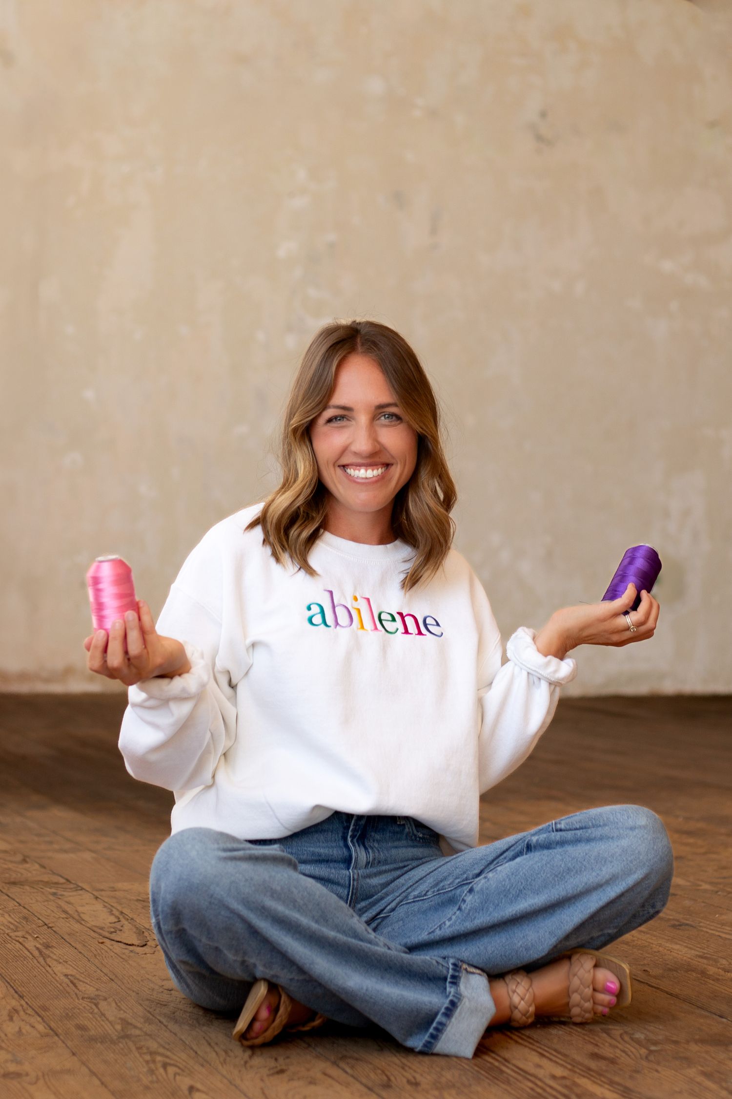 Smiling woman sitting cross-legged holding colorful thread spools wearing Abilene embroidered sweatshirt.