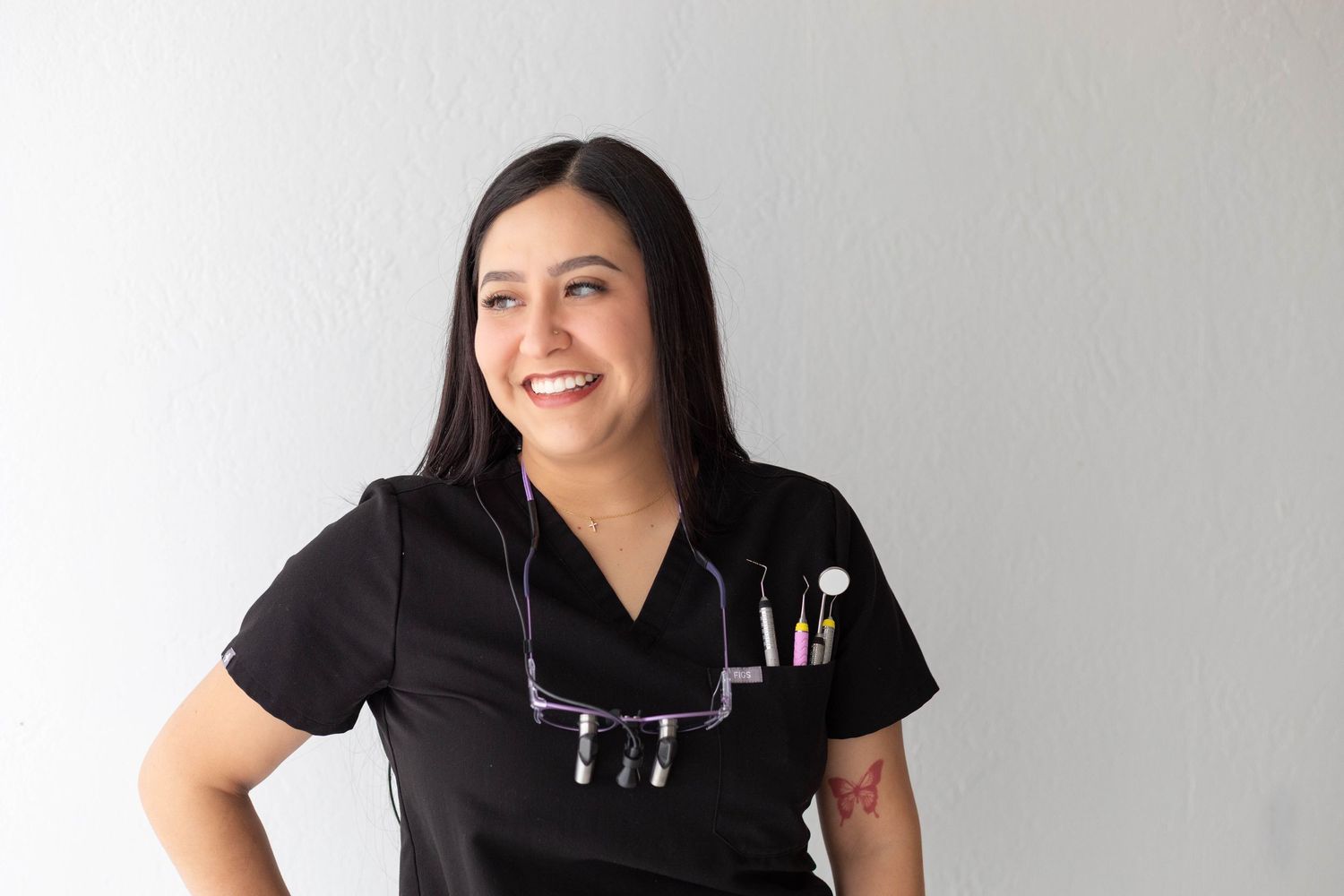 Happy female healthcare worker in black scrubs with stethoscope smiling against white wall.