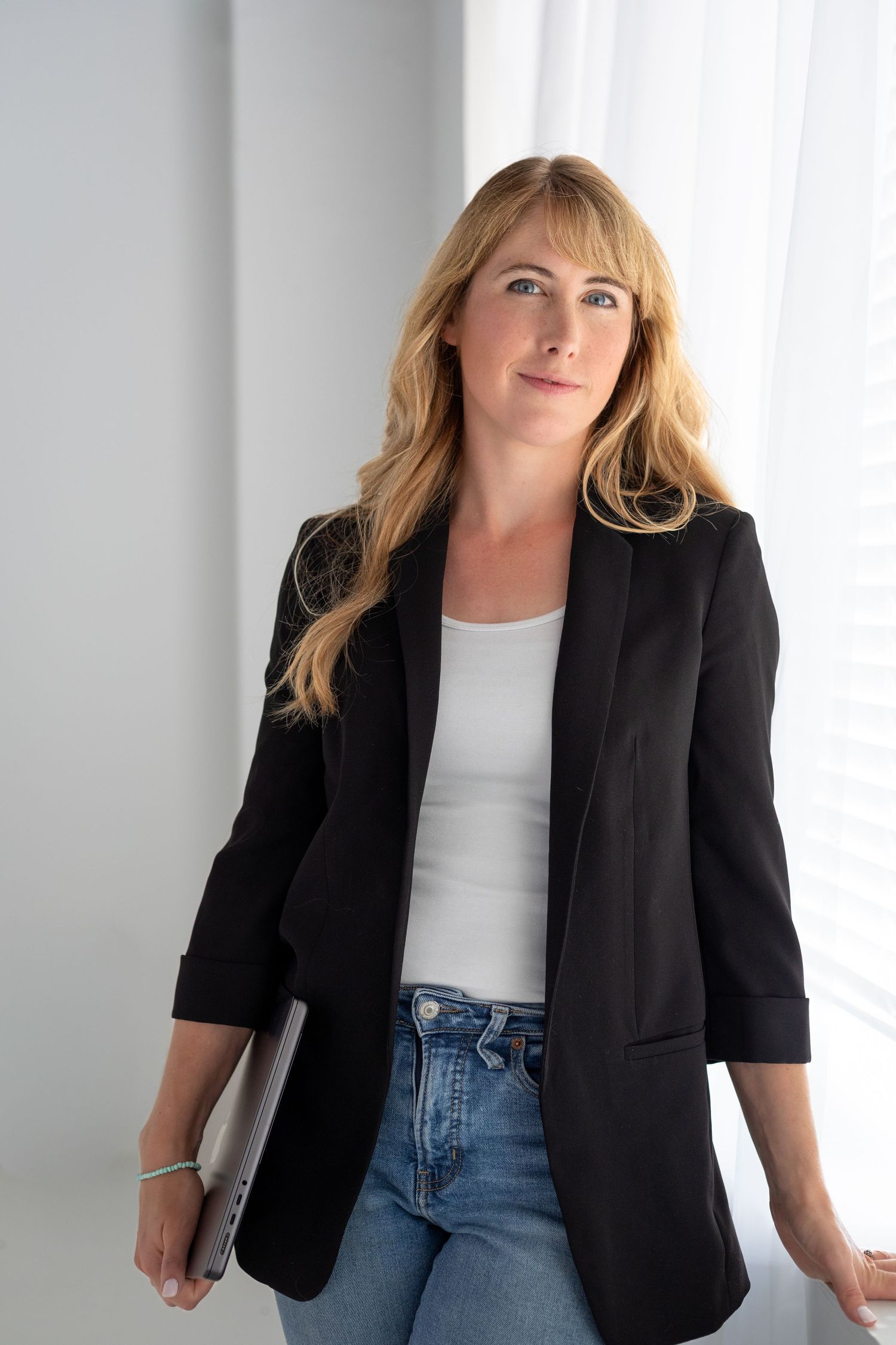 Professional blonde woman in black blazer and jeans holding laptop near bright window.