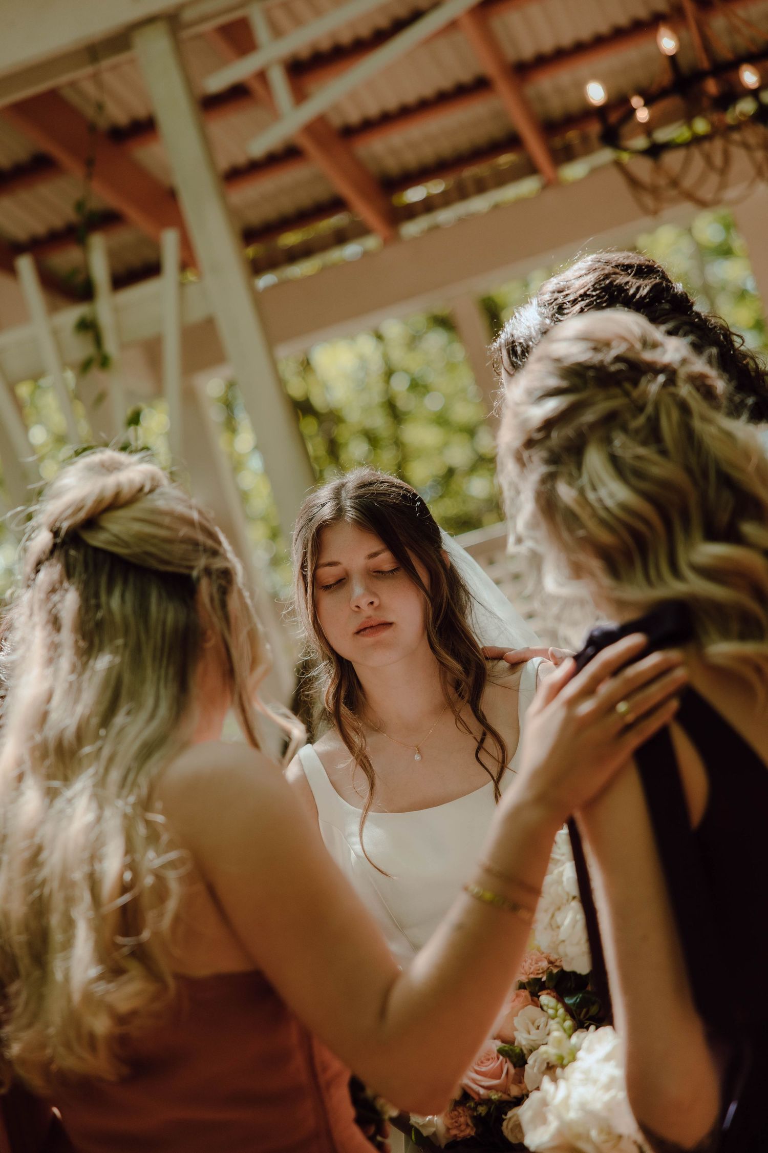 Bride in white dress stands with bridesmaids in elegant updo hairstyles at an outdoor wedding venue.