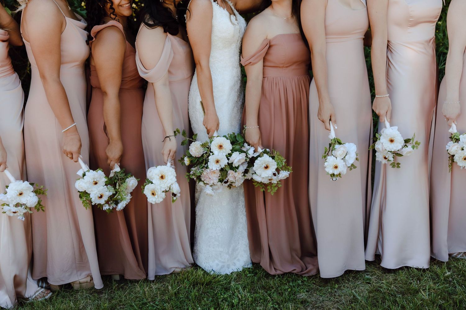 Bride and bridesmaids in blush and taupe gowns holding white floral bouquets lined up outdoors.