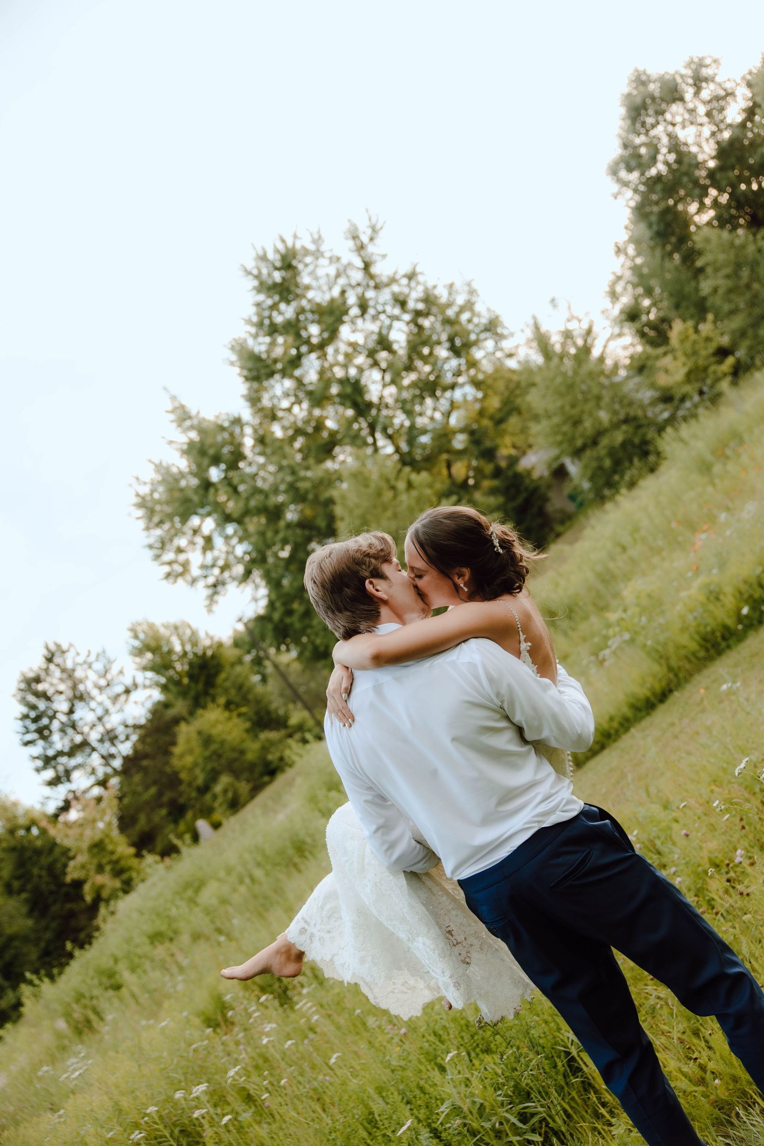 Groom lifting bride in white dress, sharing a kiss in a lush green meadow on their wedding day.