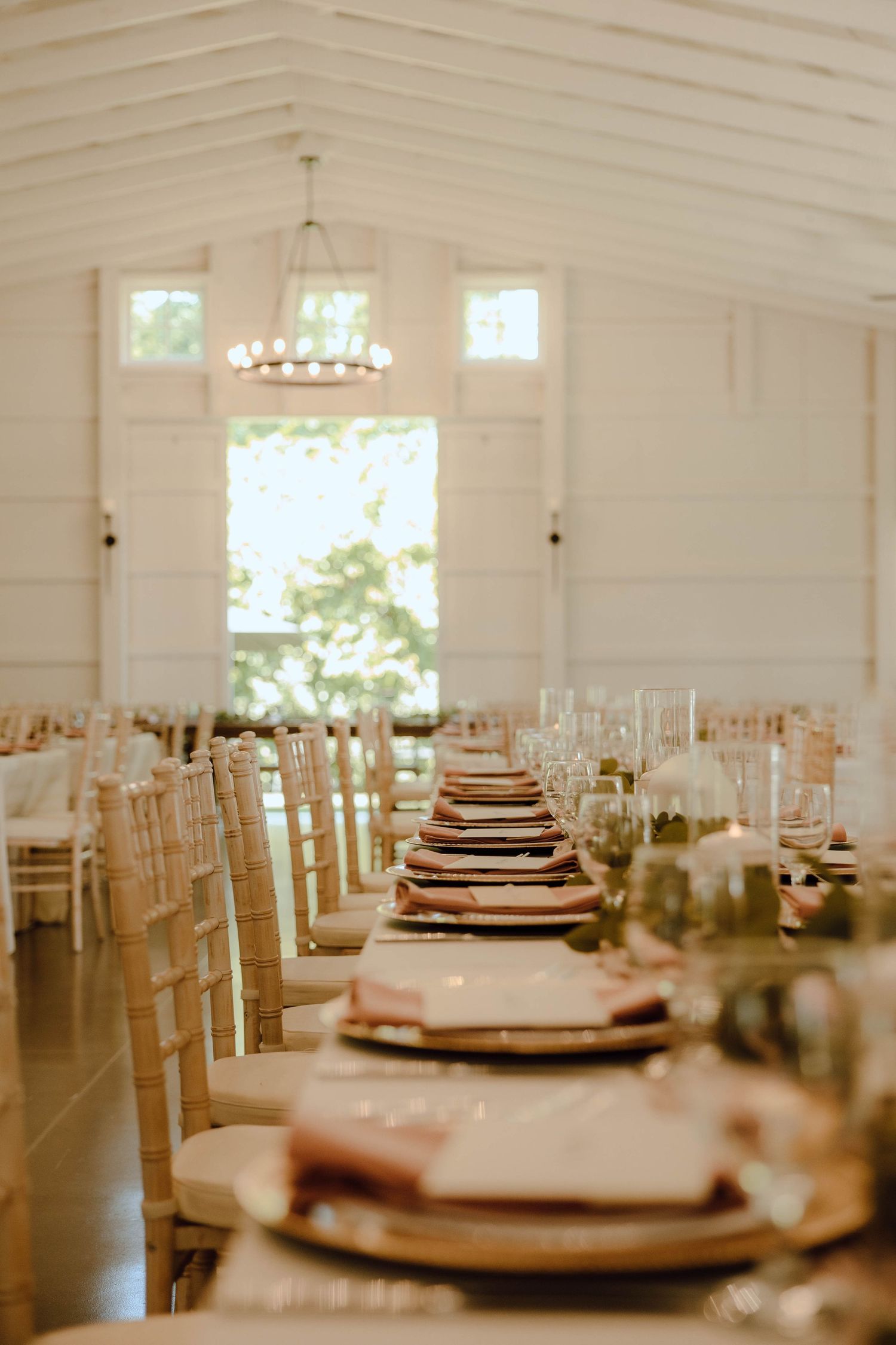 Elegant barn wedding reception with long tables, chiavari chairs, blush linens, and a chandelier in a white venue.