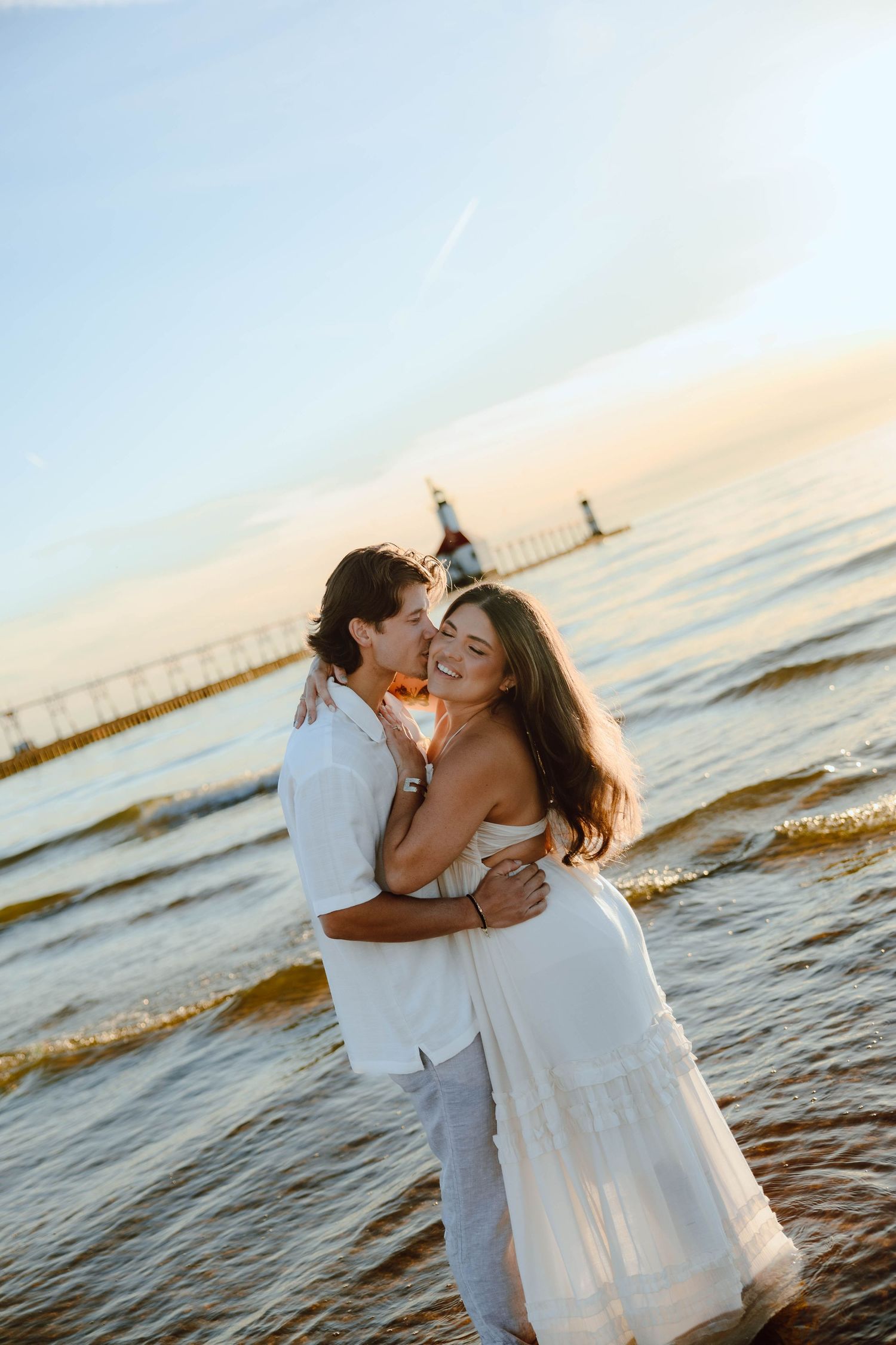 Couple embracing on a beach at sunset, woman in white dress smiling as man kisses her cheek near a pier.