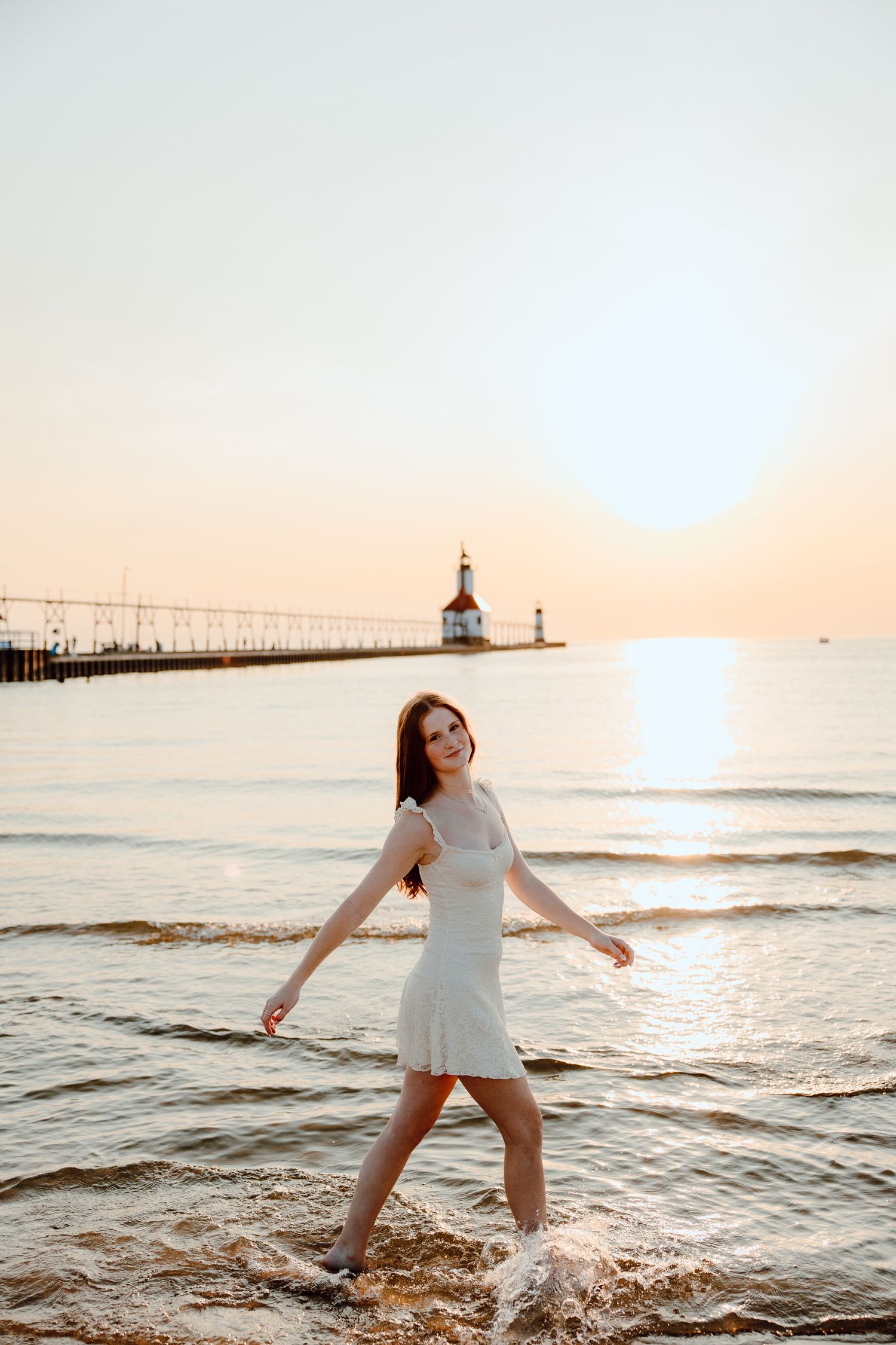 Someone in a white dress splashes in the ocean waves at sunset with a lighthouse in the background.