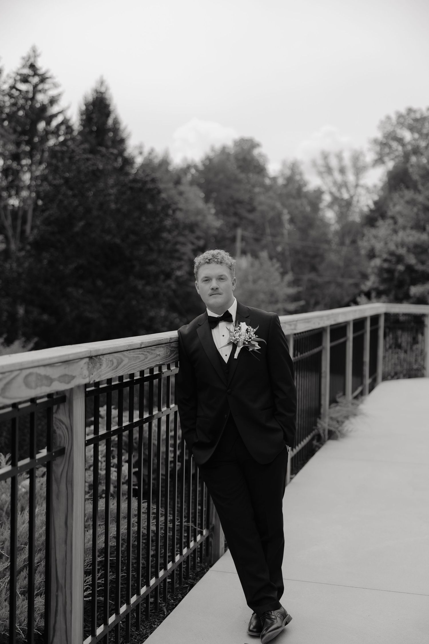 Groom in black tuxedo with boutonniere leaning on bridge railing outdoors in black and white wedding photo.