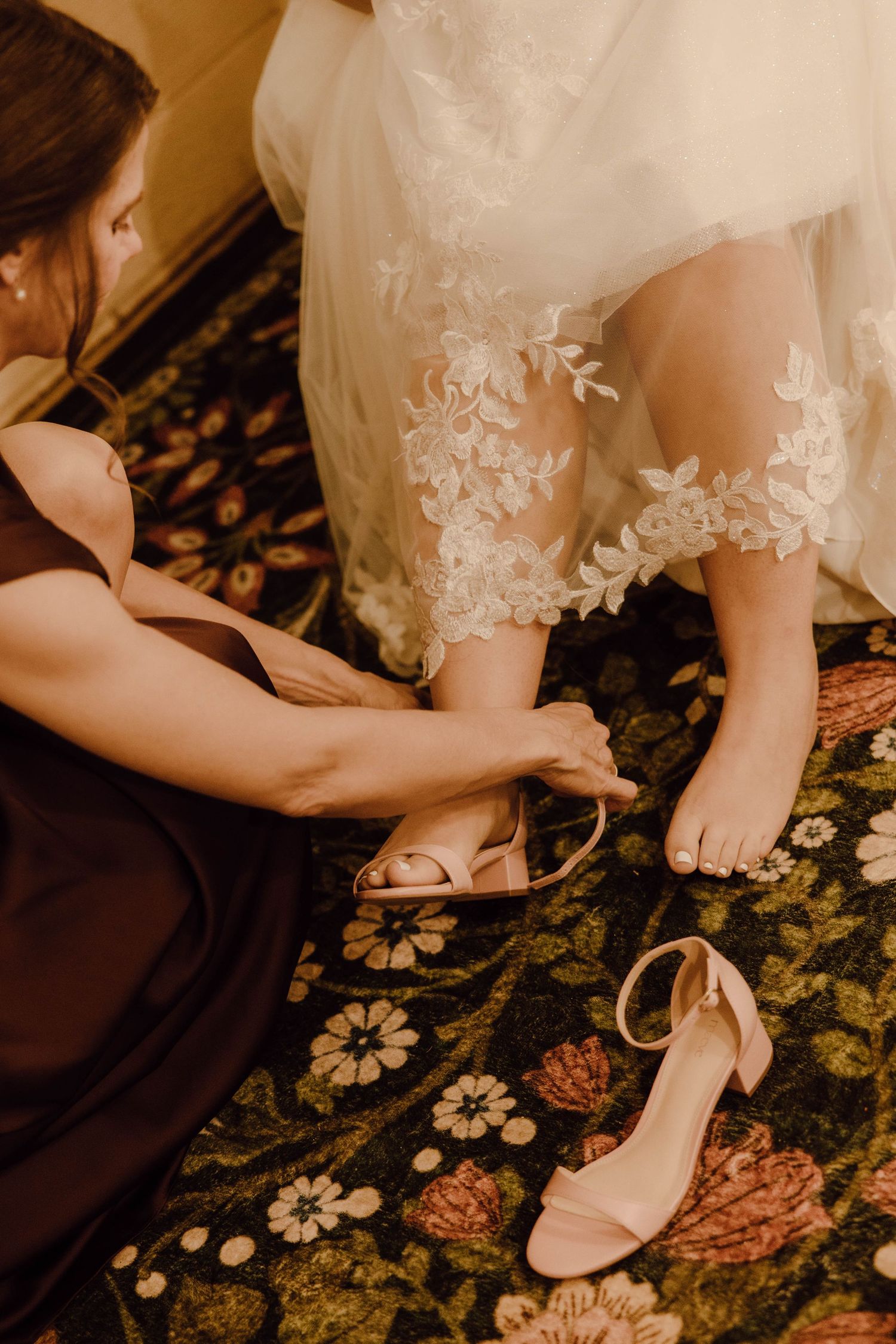 Bridesmaid helping bride put on nude heeled sandals before wedding ceremony on floral carpet.