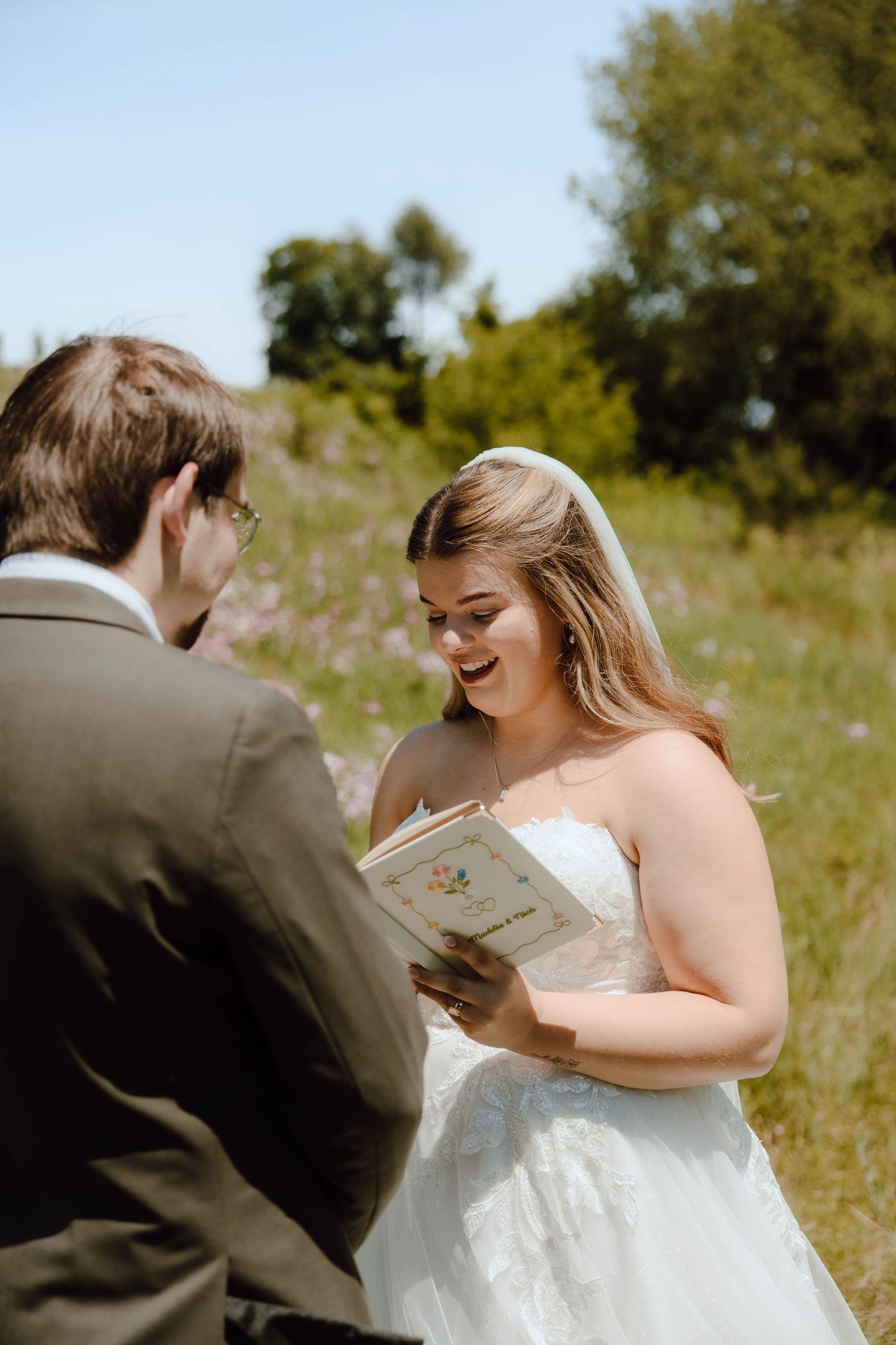Smiling bride in white strapless gown reads vows from a floral book outdoors on a sunny wedding day.