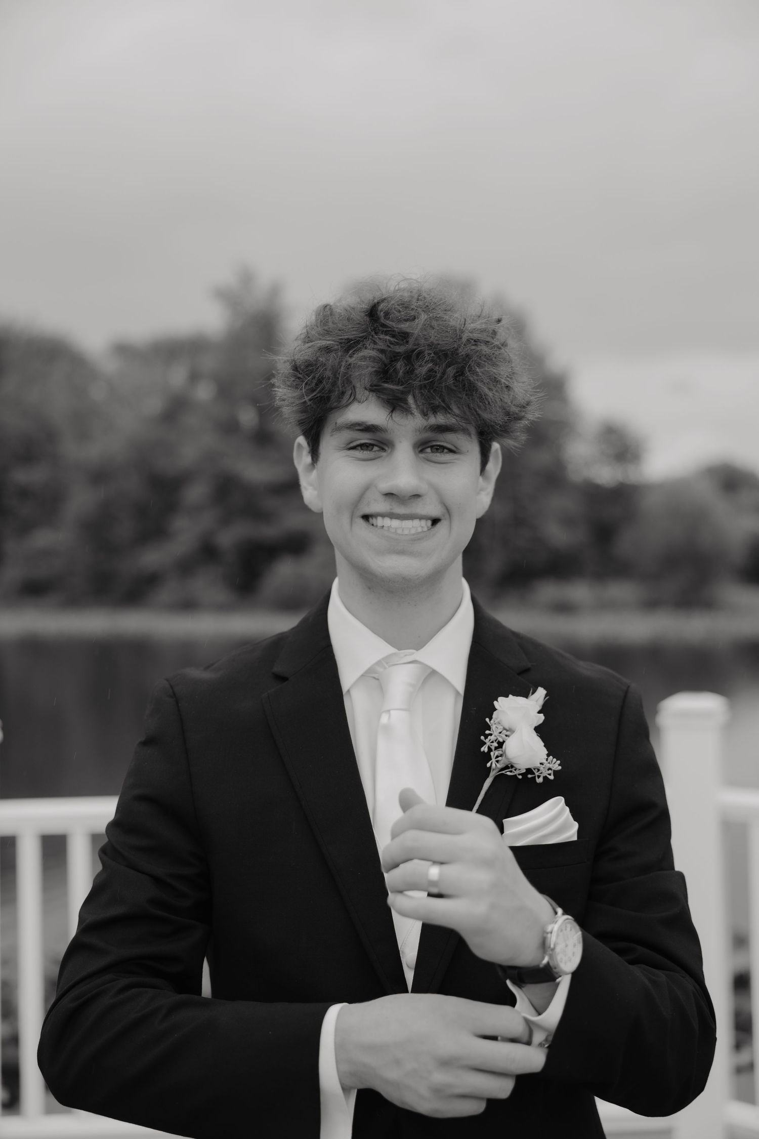 Black and white portrait of smiling groom in suit holding boutonniere by lakeside.