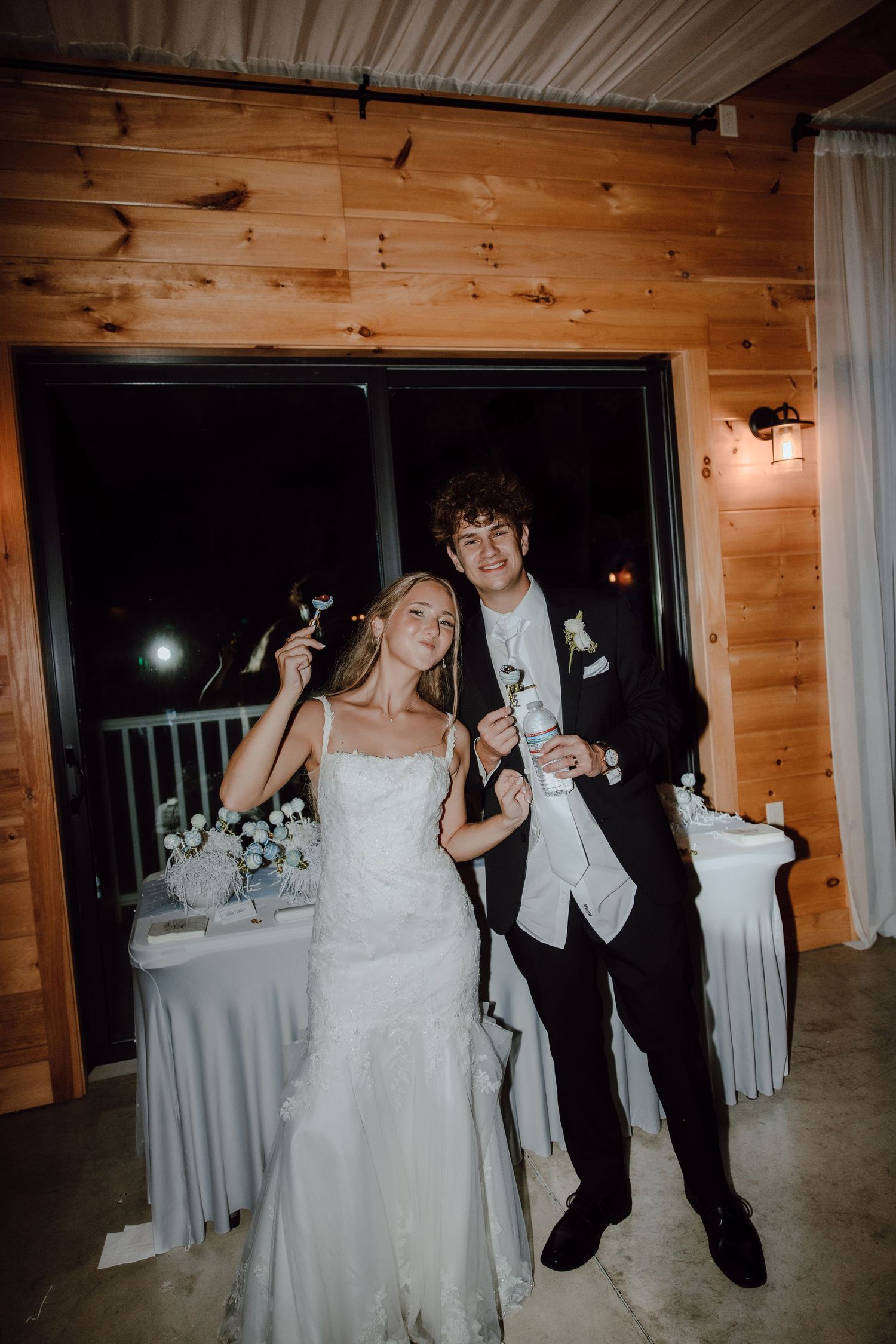 Smiling newlywed couple poses playfully with cake cutting set at indoor rustic wood-paneled reception venue.