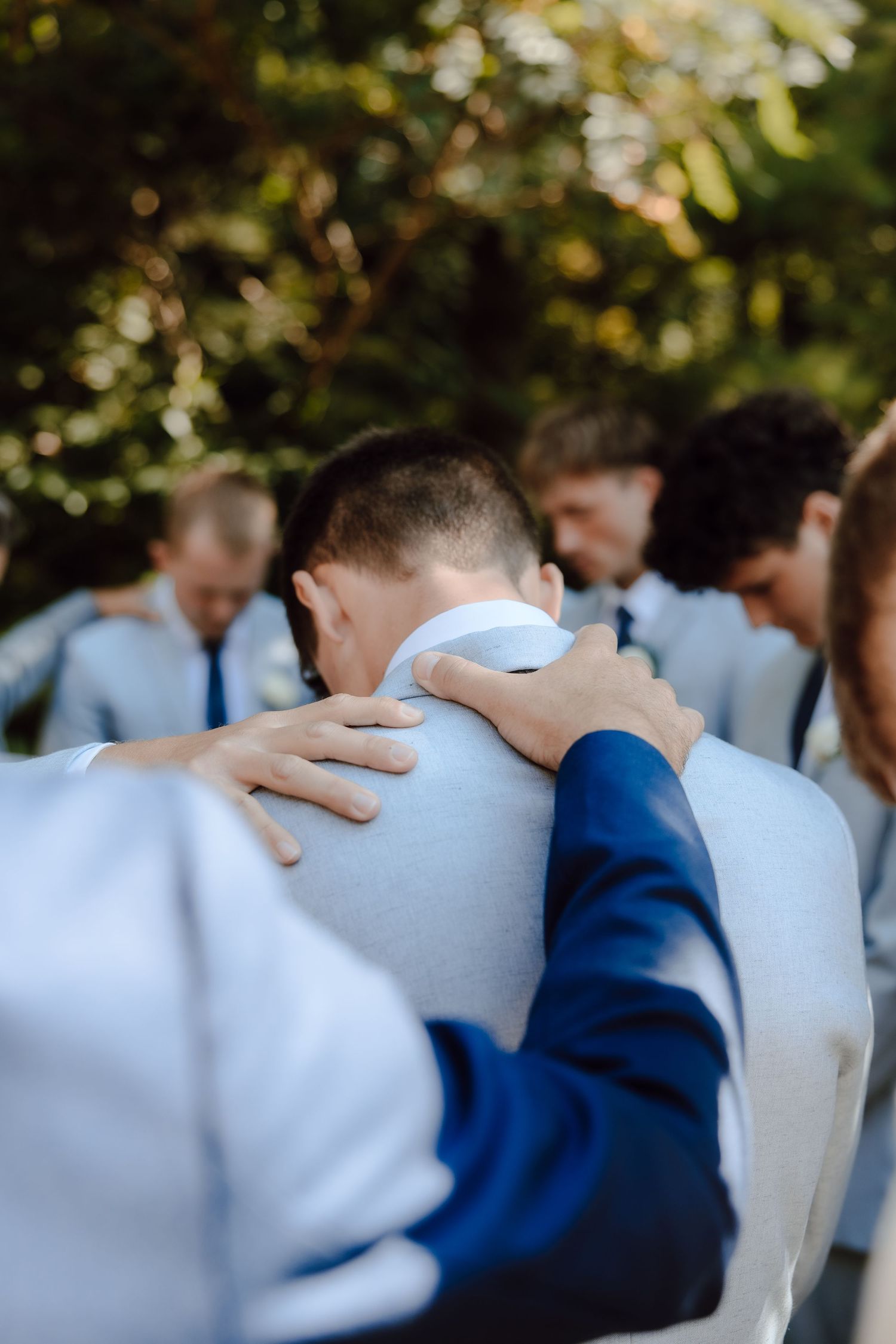 Close-up of groomsmen placing hands on grooms shoulders in prayer before wedding ceremony outdoors.
