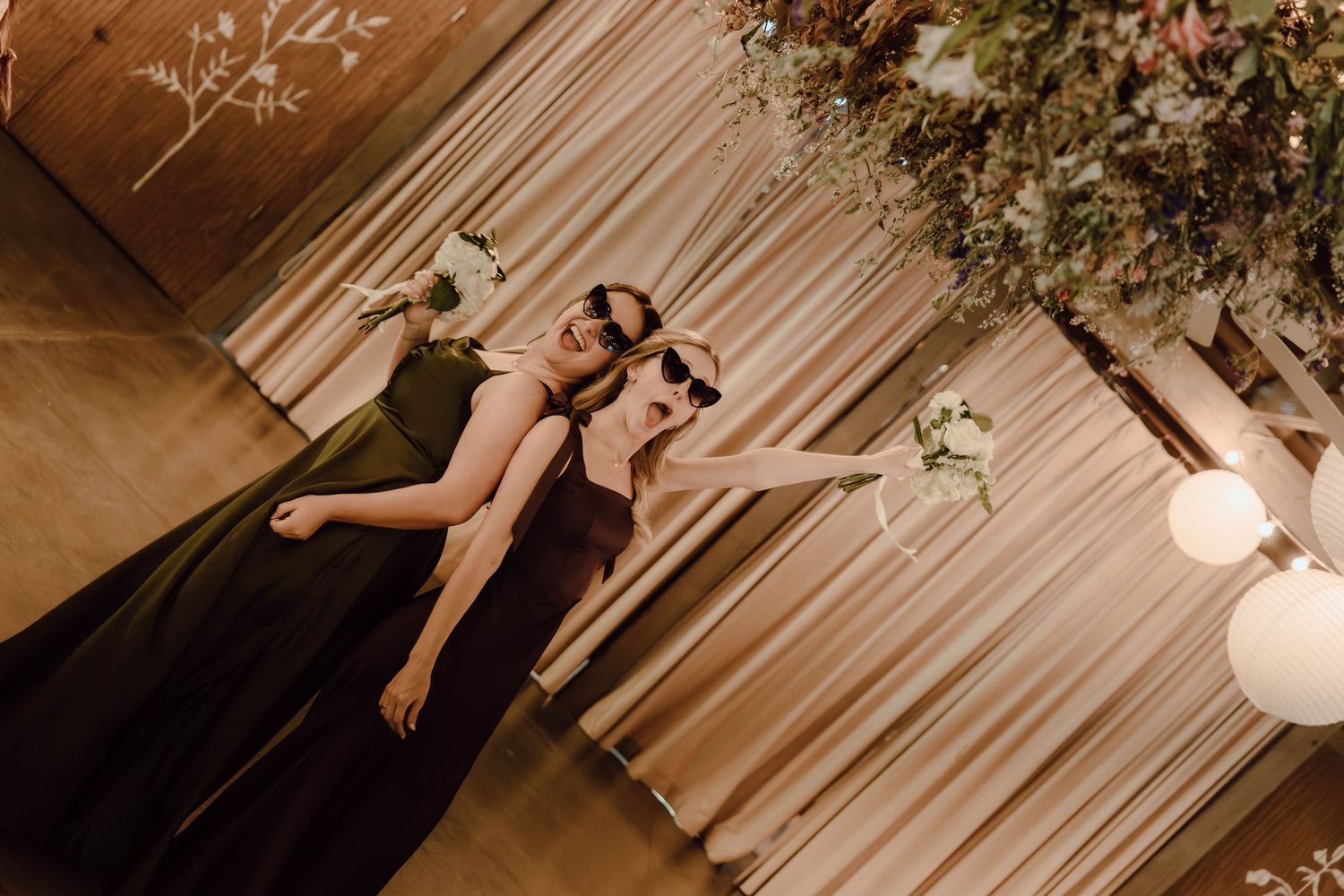 Two bridesmaids in black gowns holding bouquets near floral arrangements and draped curtains at a wedding reception.