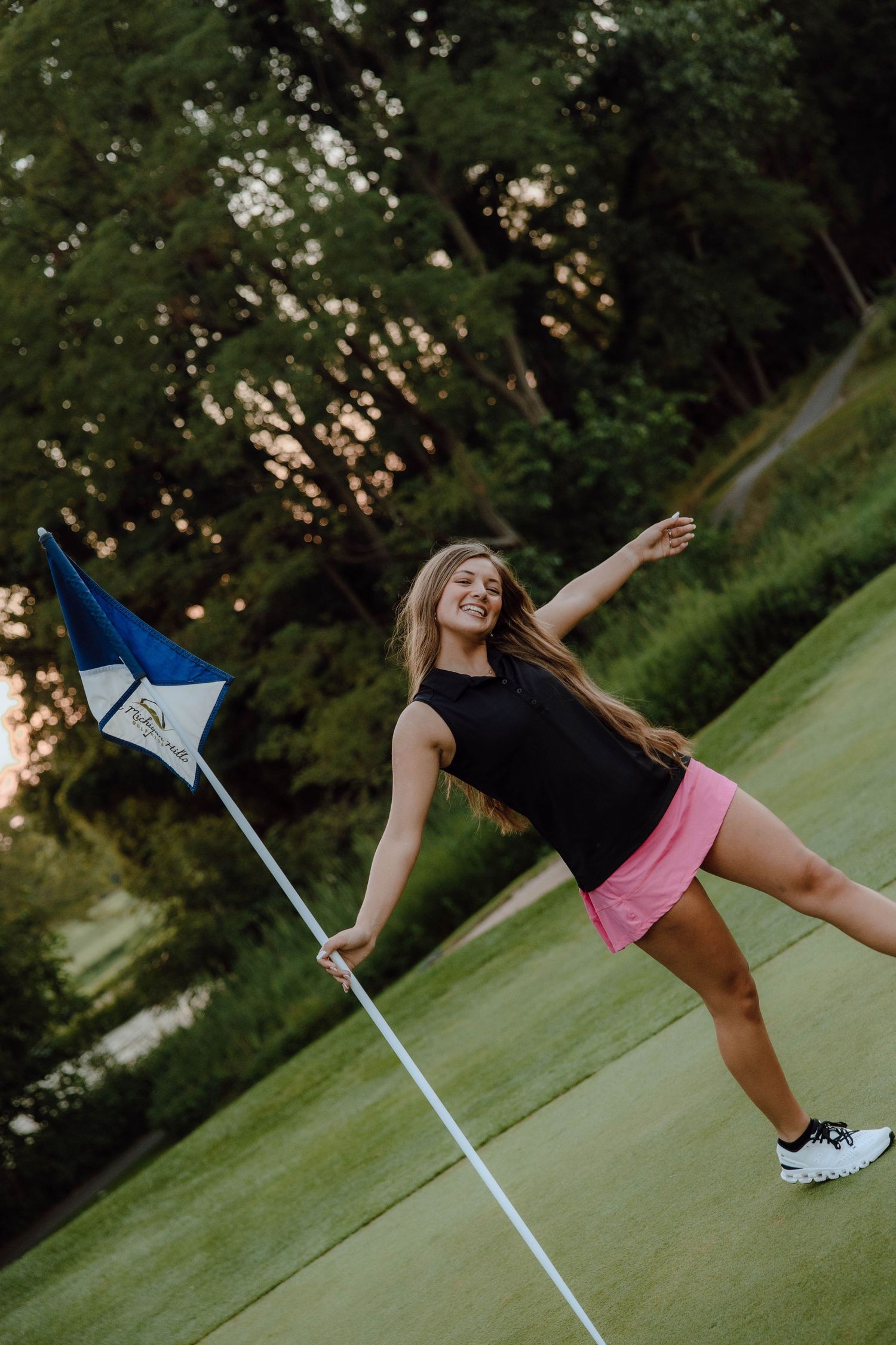 Young woman in pink skirt holding golf flag on green course, posing playfully at sunset.