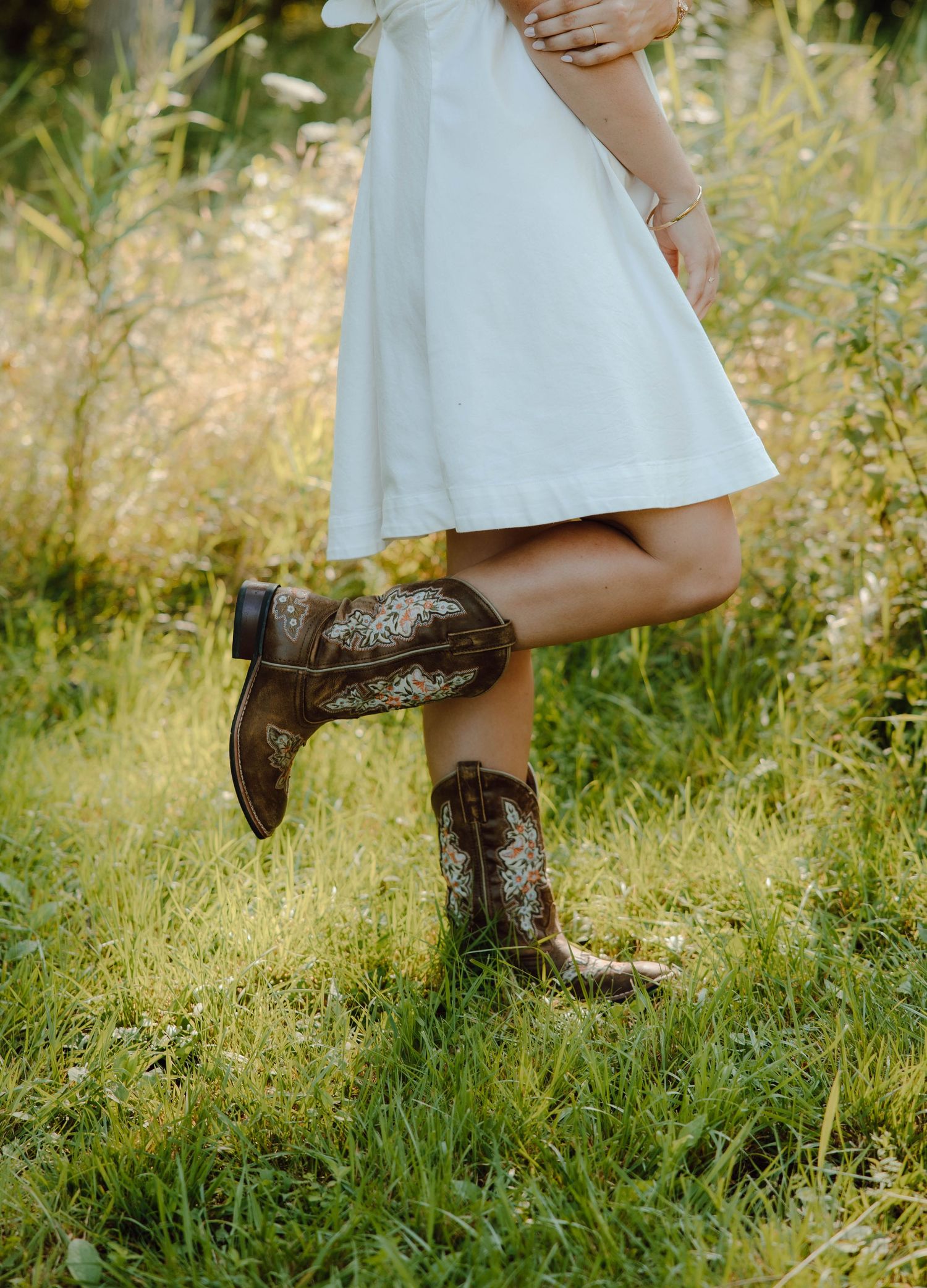 Woman in white dress and brown cowboy boots with floral embroidery standing on green grass in a natural outdoor setting.