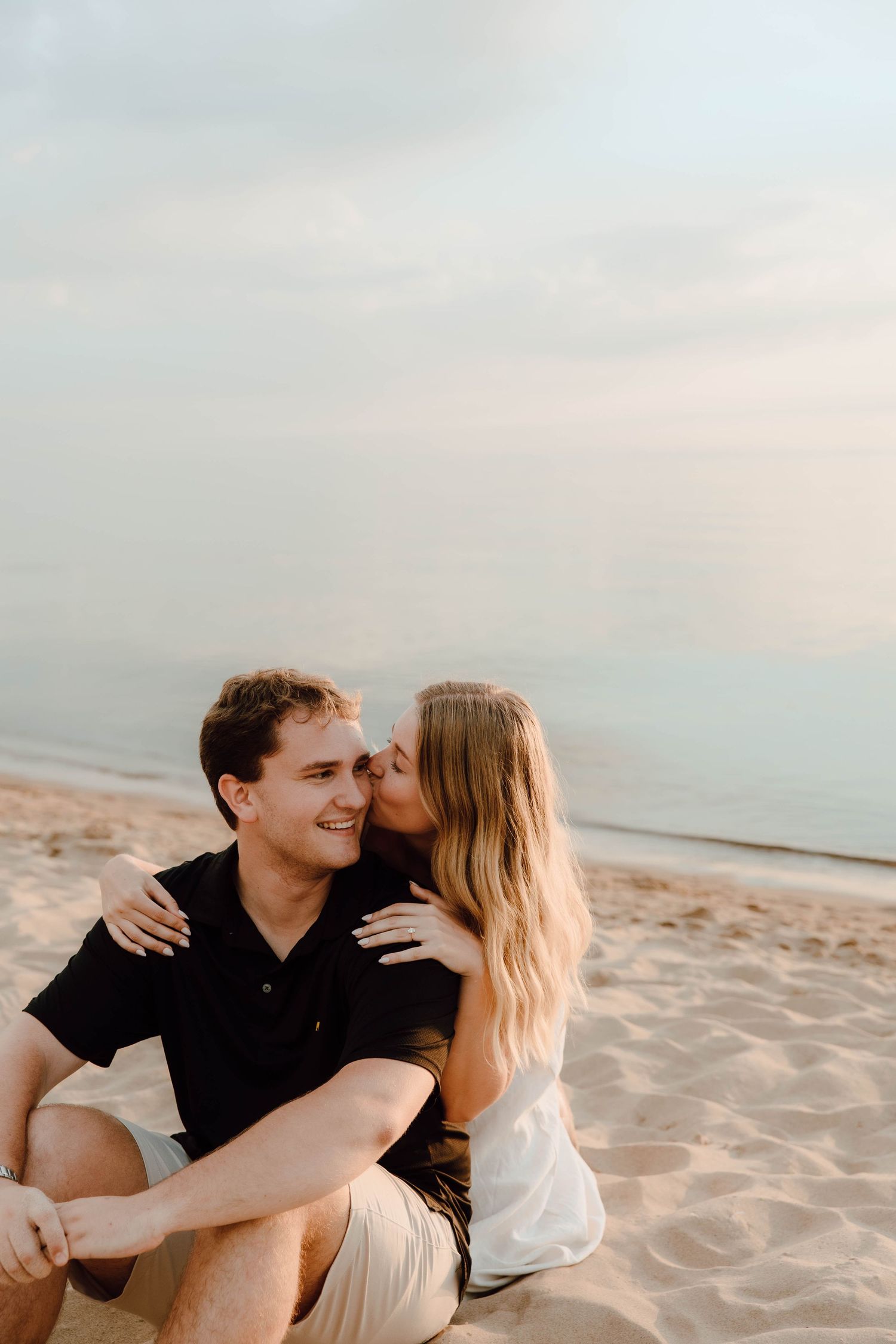 Couple sitting on sandy beach at sunset, woman kissing man on cheek while he smiles, ocean in background.