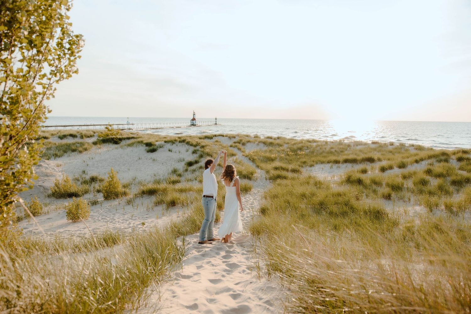 Couple walking hand in hand on a sandy beach path toward the ocean at sunset near a lighthouse.