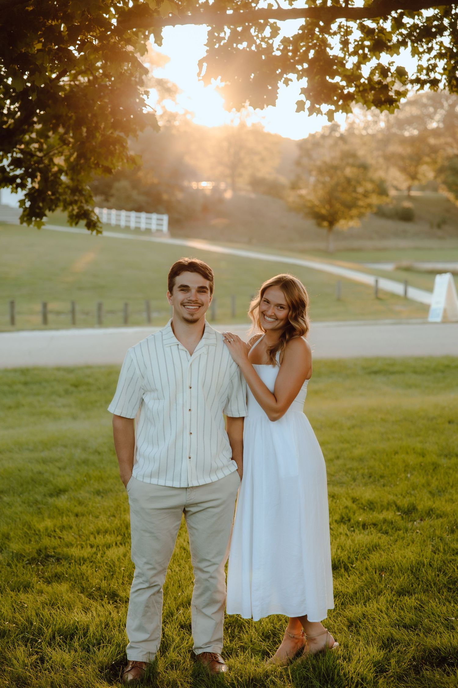 A couple poses at golden hour on a lush green lawn, man in striped shirt and woman in white dress, with trees and fence behind.