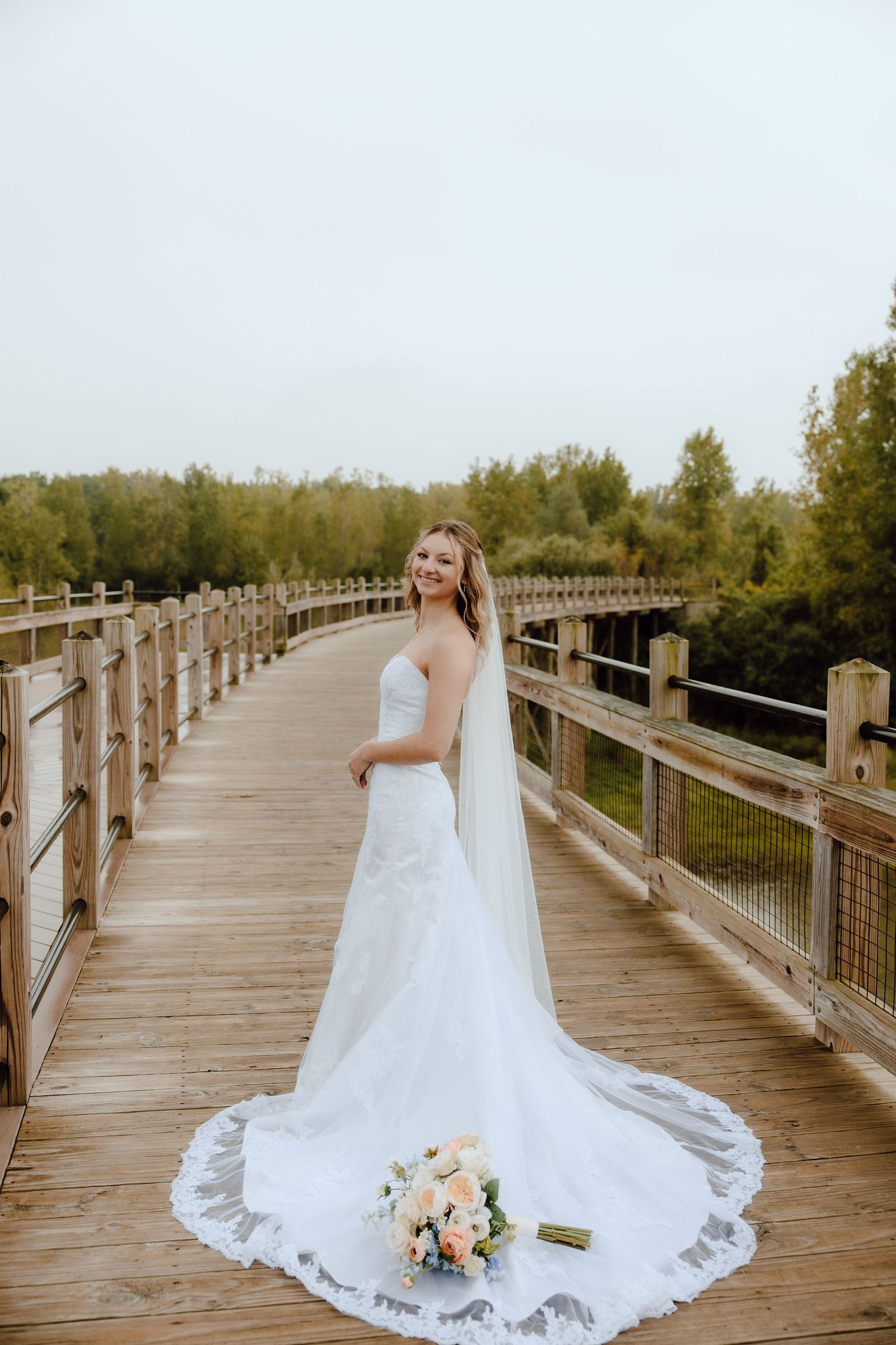 Bride in a white strapless gown with long veil holds bouquet on a wooden bridge surrounded by trees.