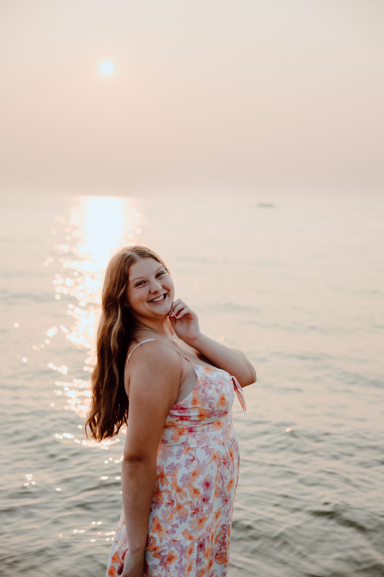 Person in floral dress poses by ocean during sunset with glowing reflection on water.