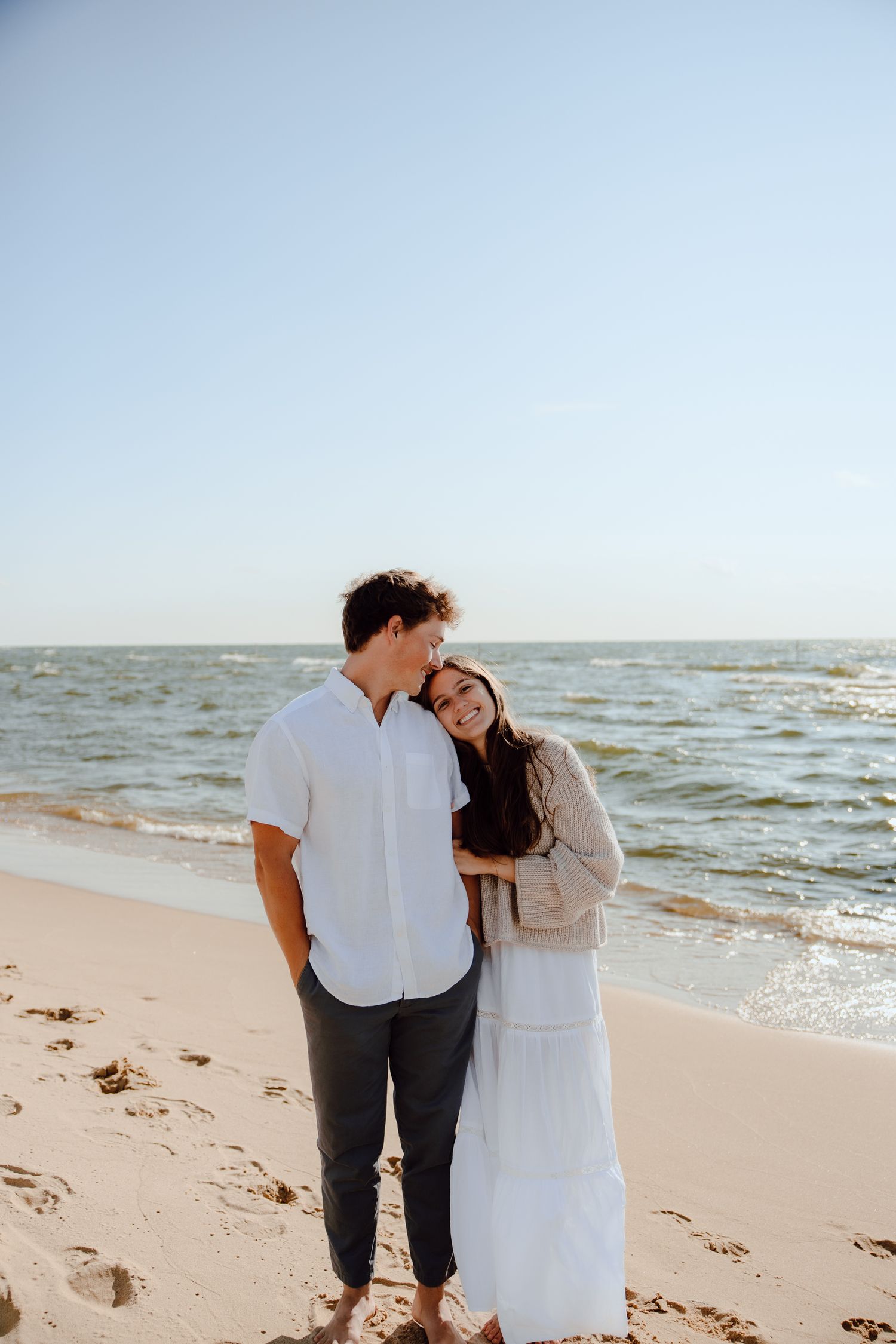 Couple in white clothing embracing on beach with ocean waves in background.