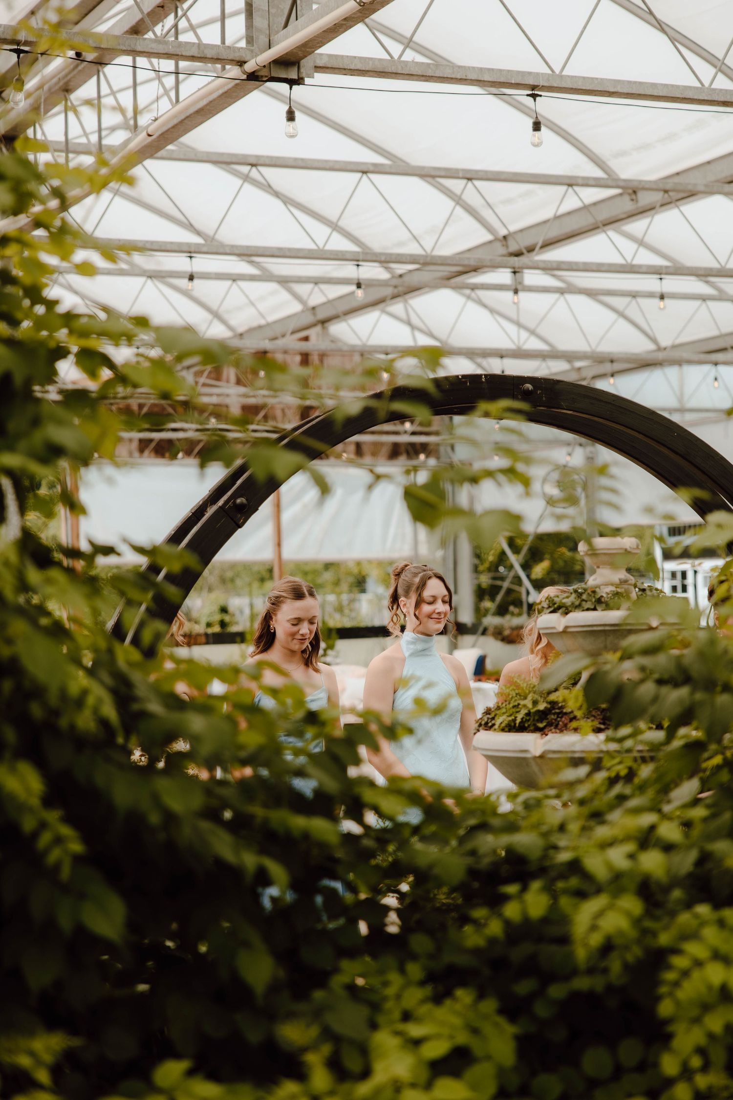 Two bridesmaids in light blue dresses laugh together inside a greenhouse wedding venue.