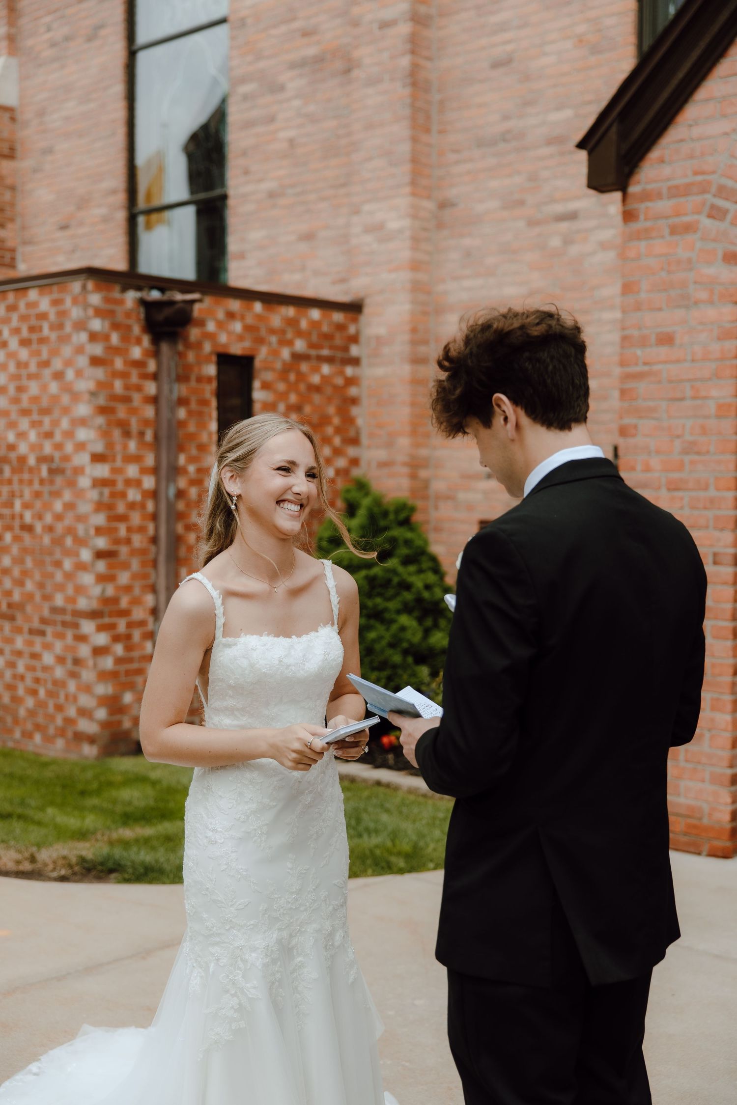 Smiling bride in lace wedding dress exchanging vows with groom outside brick building.