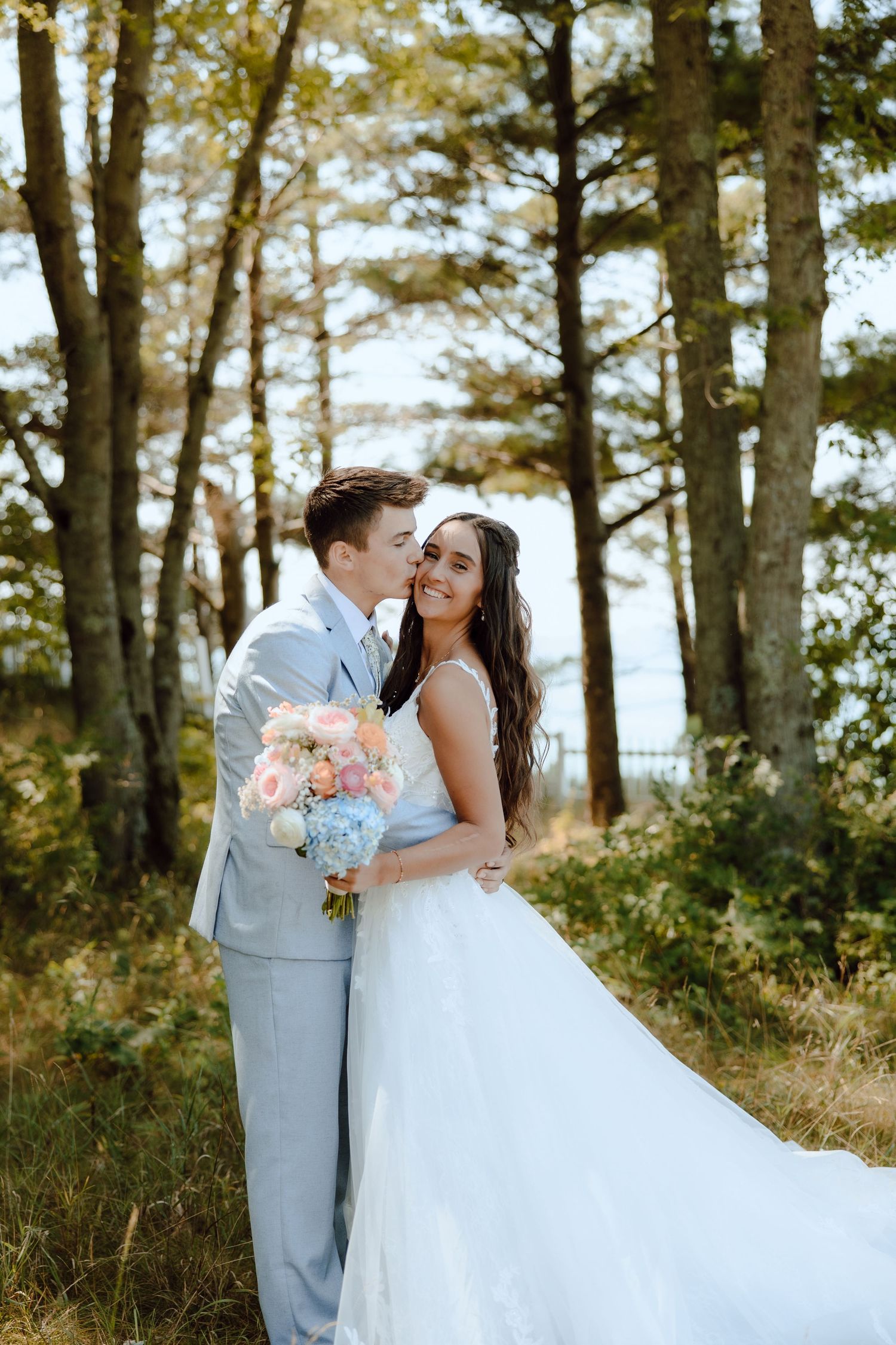 Bride and groom embracing in wooded setting, bride holding pink and blue bouquet, groom in gray suit.