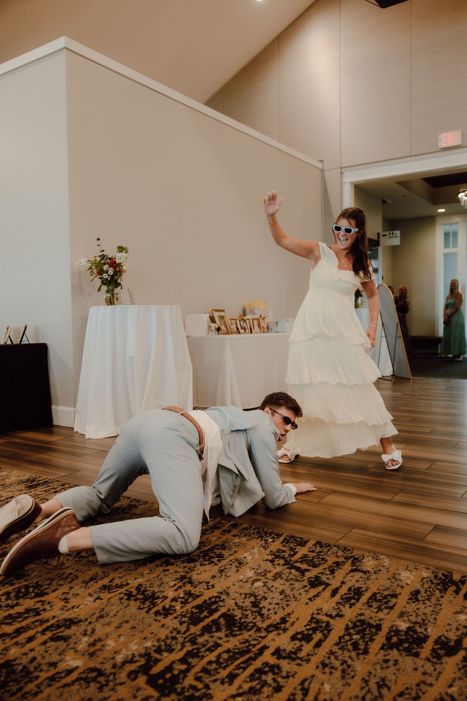 Bride and groom dancing playfully at wedding reception, groom crawling on floor while bride raises hand wearing sunglasses.