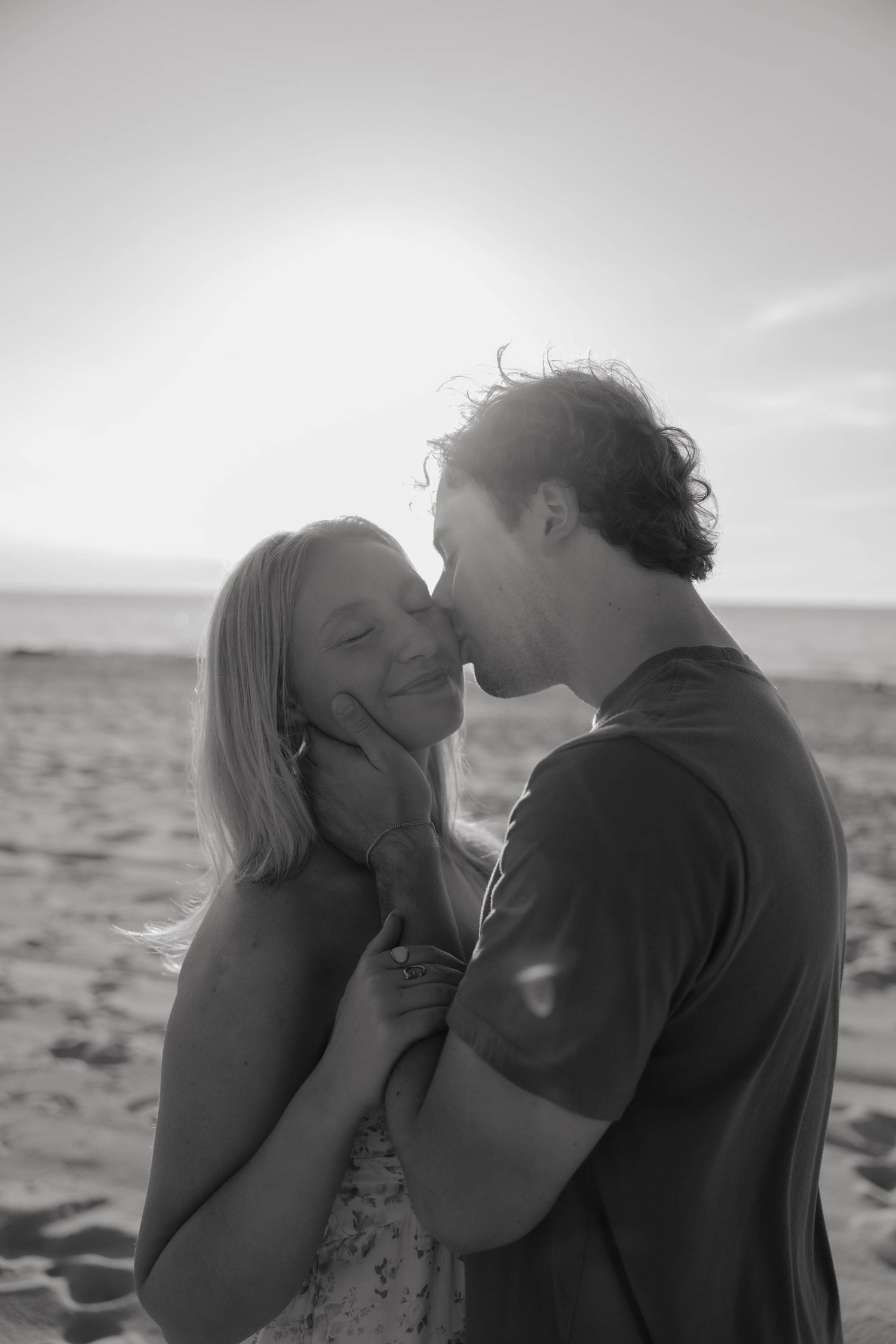Black and white photo of a man kissing a smiling woman on a beach.