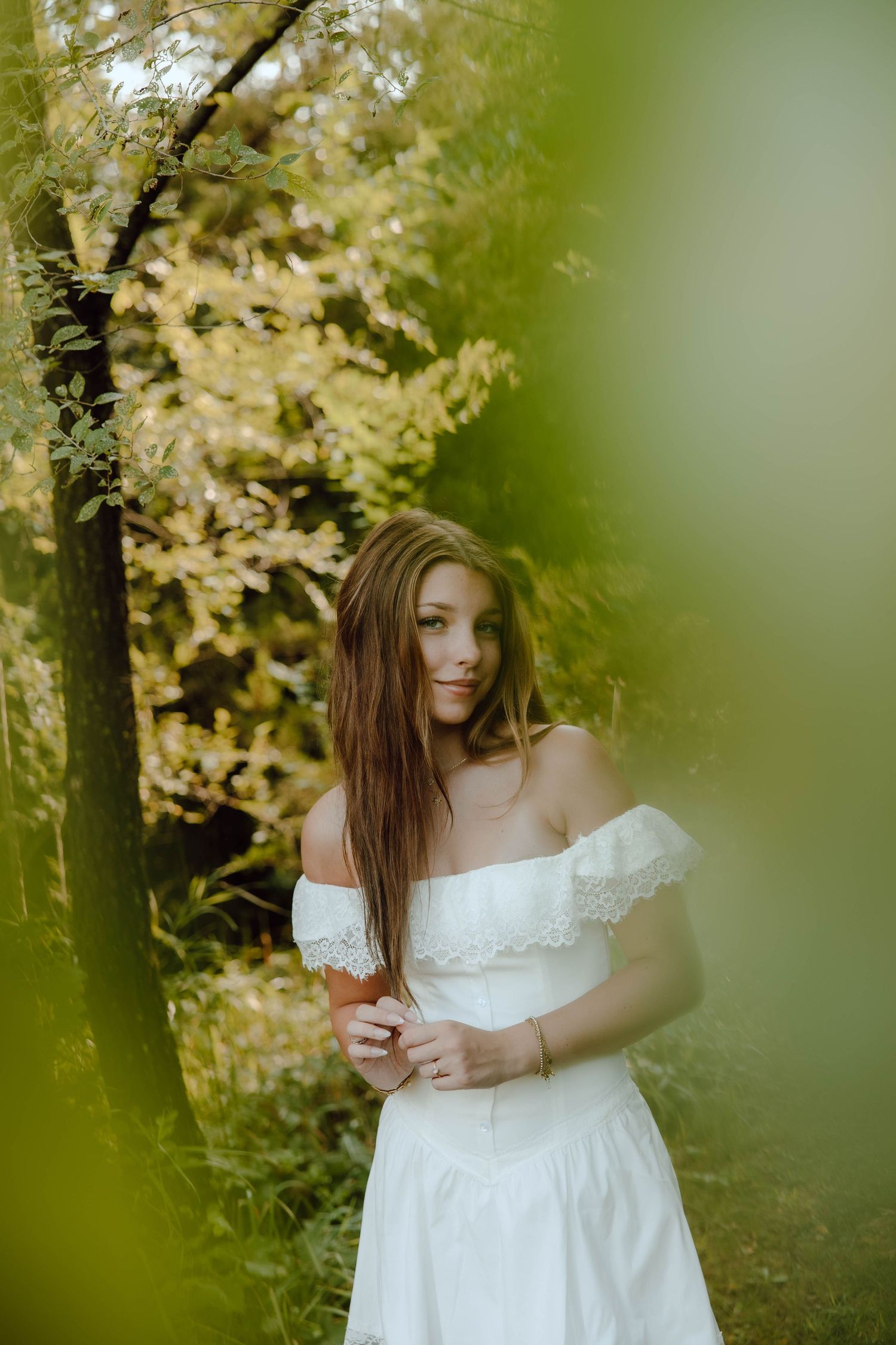 Young woman in white off-shoulder dress standing among golden autumn trees in a lush garden setting.