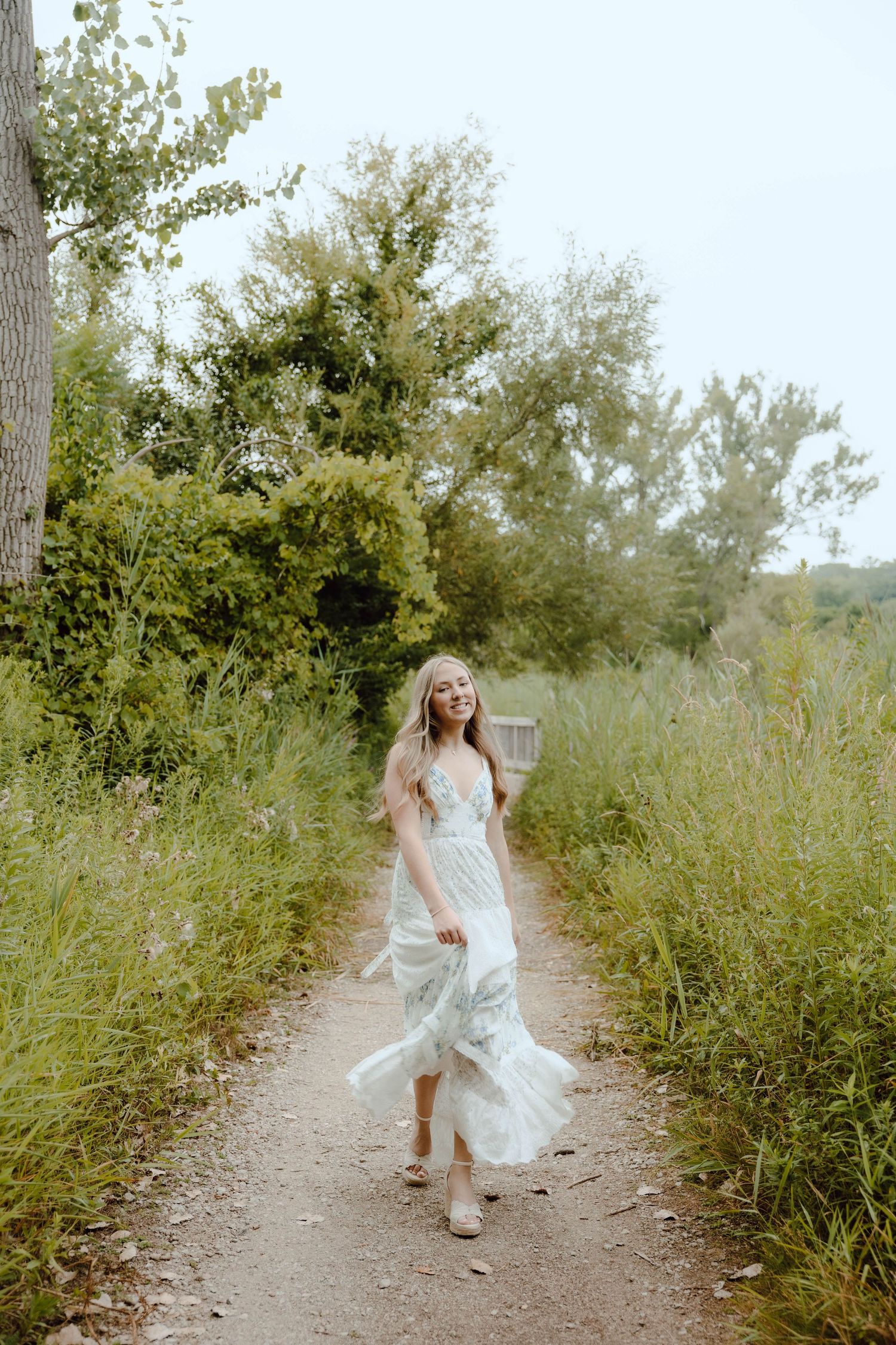 Young woman in flowing white floral dress walking on a nature path surrounded by lush green trees.