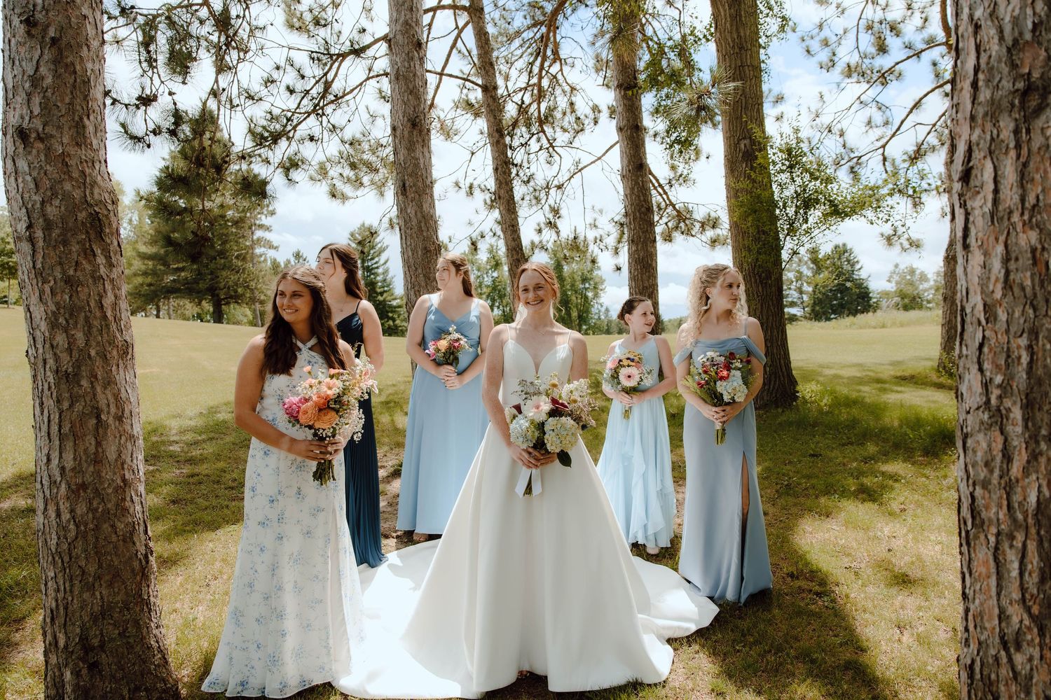 Bride in white gown poses with bridesmaids in blue dresses holding colorful bouquets among pine trees outdoors.