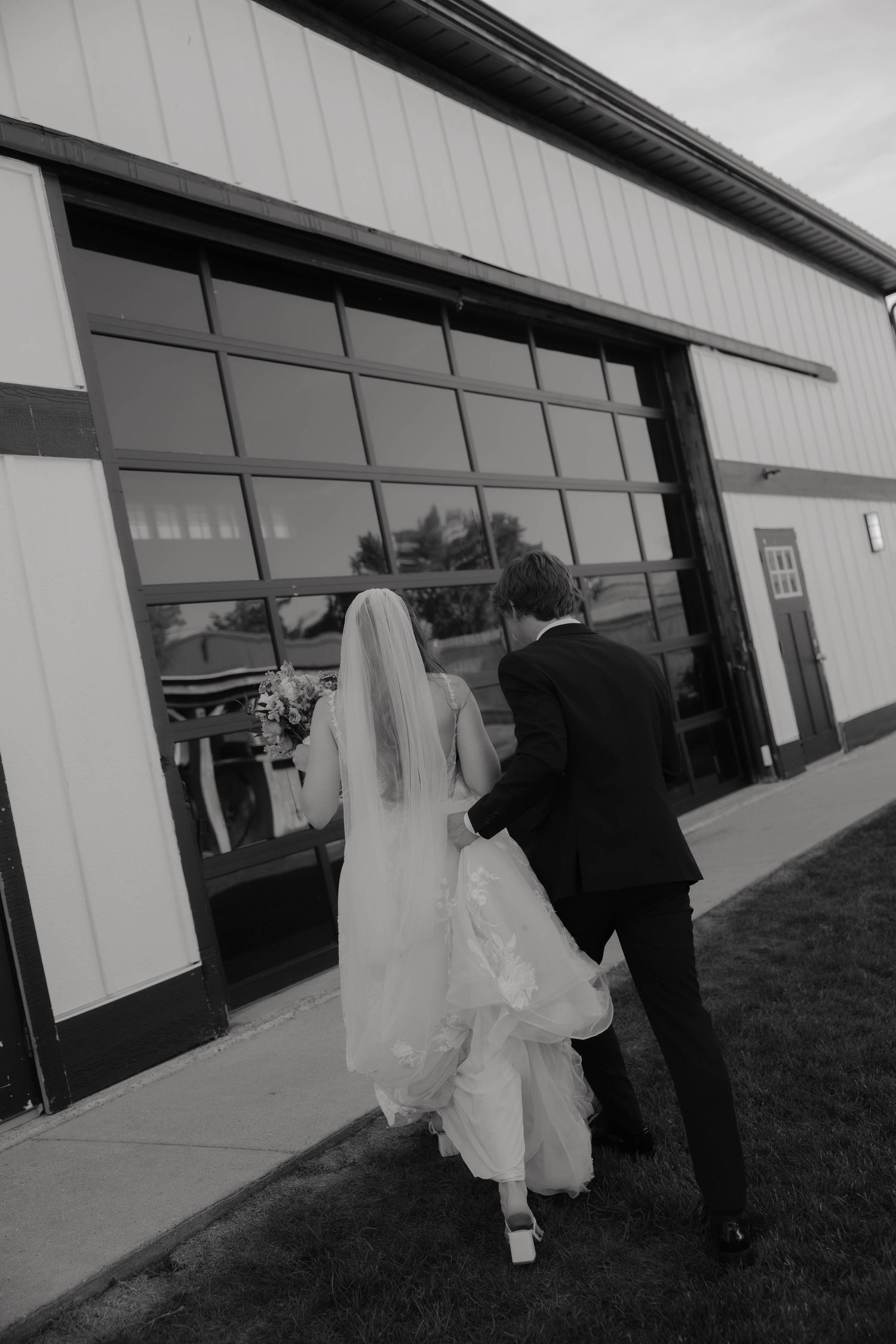 Black and white photo of bride and groom walking hand in hand toward a modern barn wedding venue.