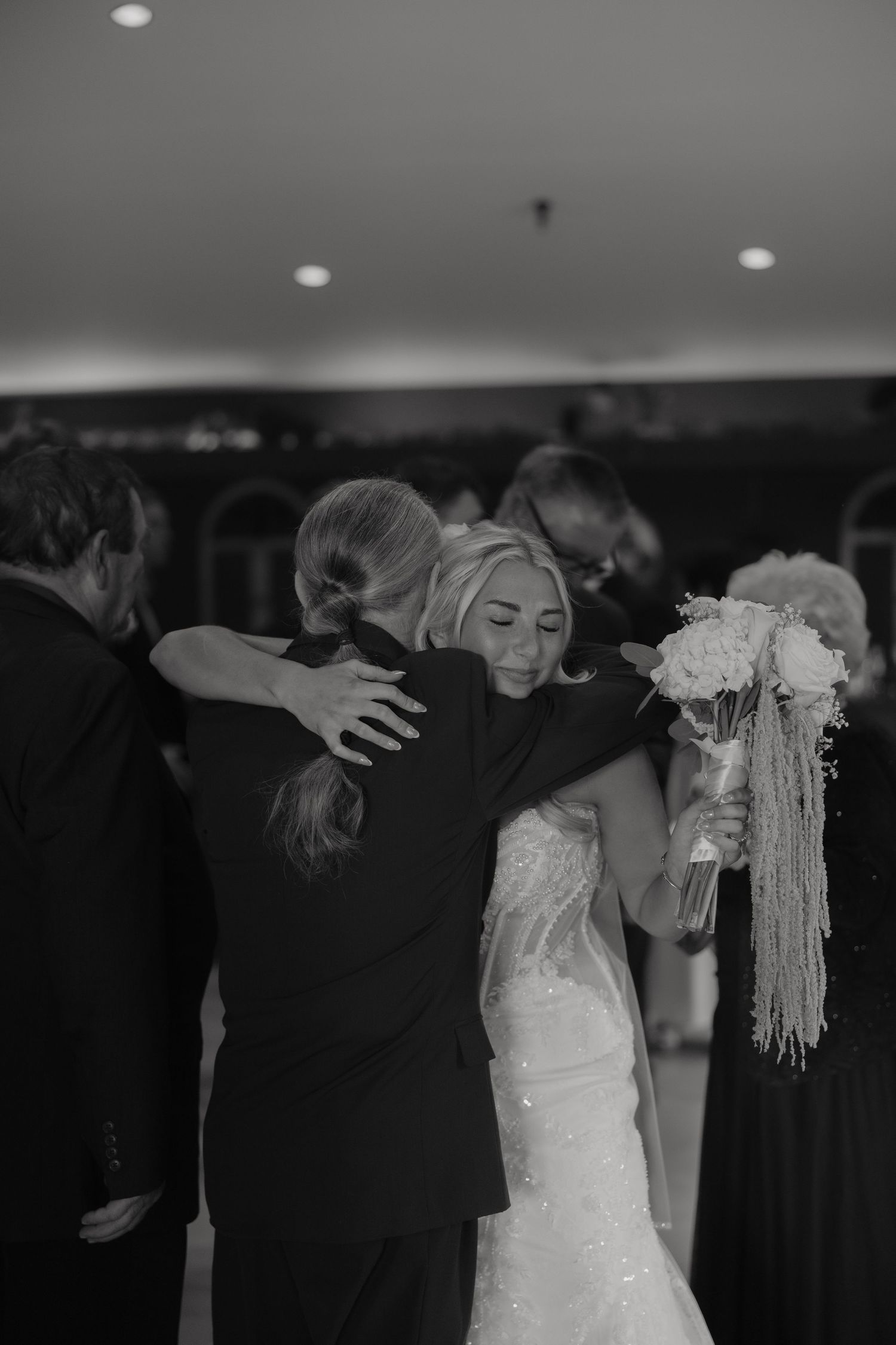Black and white photo of a bride in a lace gown hugging a guest while holding a bouquet at her wedding reception.