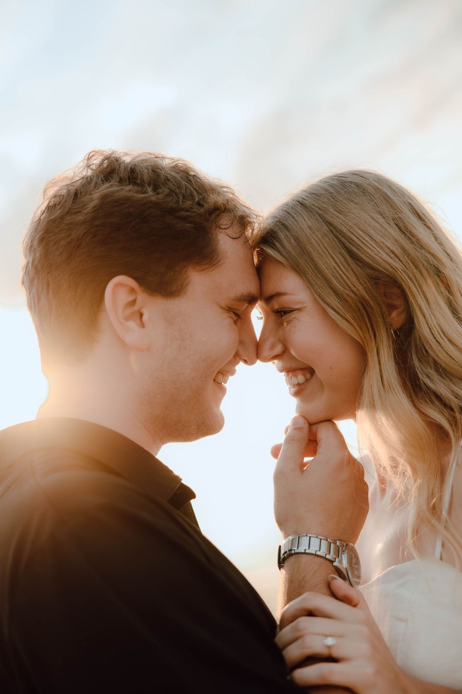 A smiling couple touching foreheads in golden sunlight, sharing an intimate romantic moment outdoors.